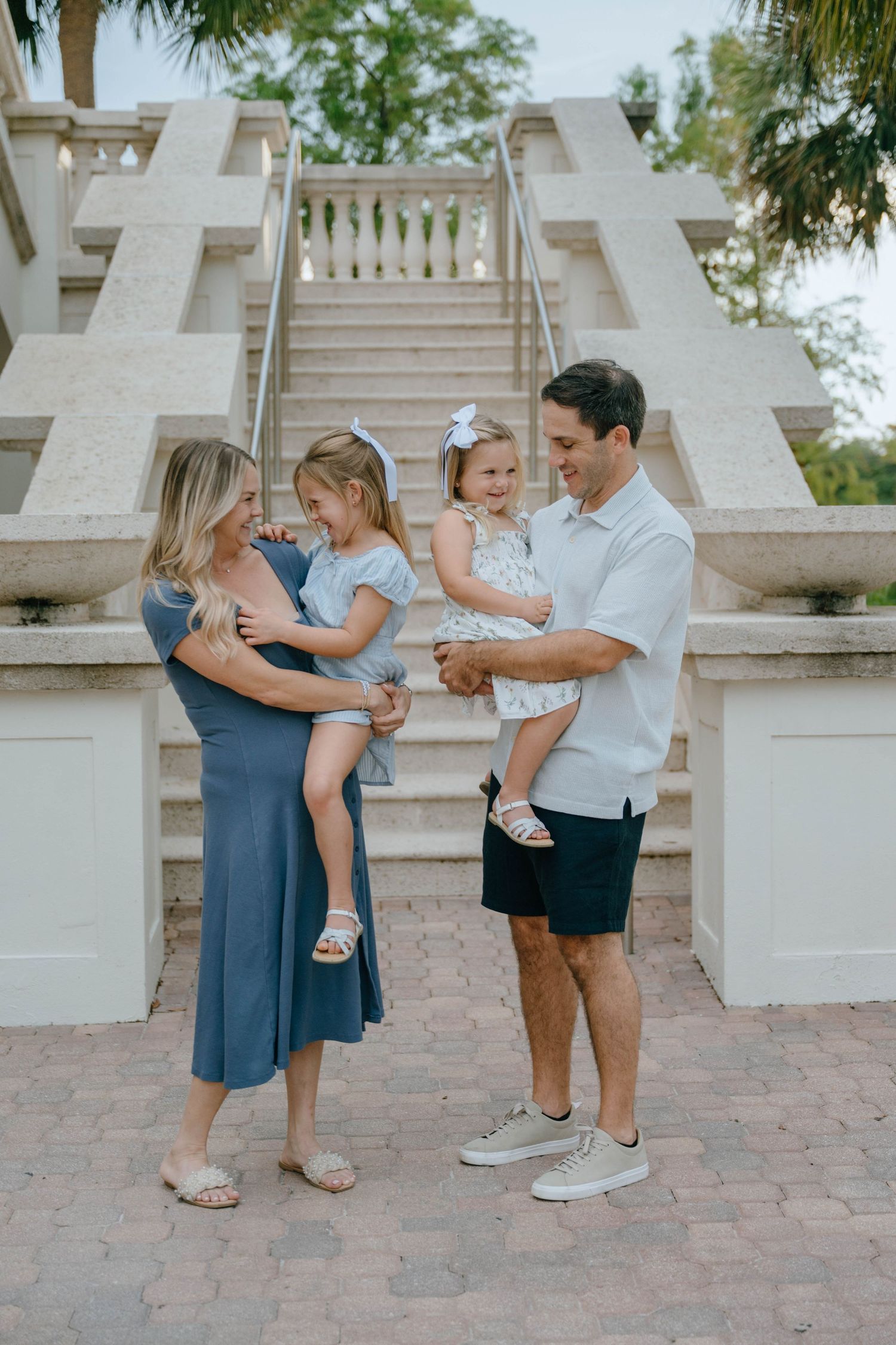 Young family with two toddler girls in blue dresses enjoy playful moment at elegant outdoor staircase.