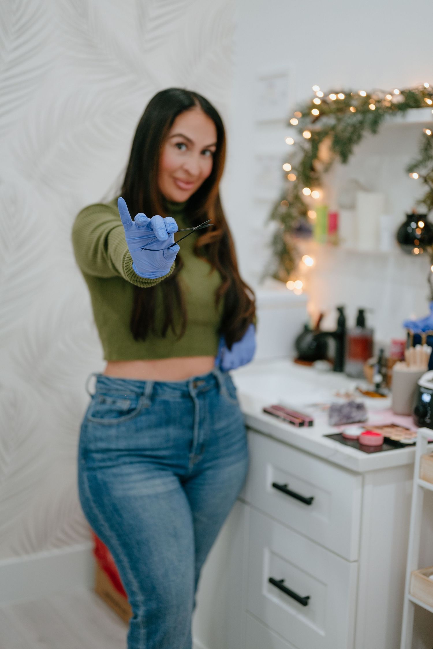 Smiling female esthetician in green crop top wearing blue gloves holds tweezers toward camera at beauty workstation.