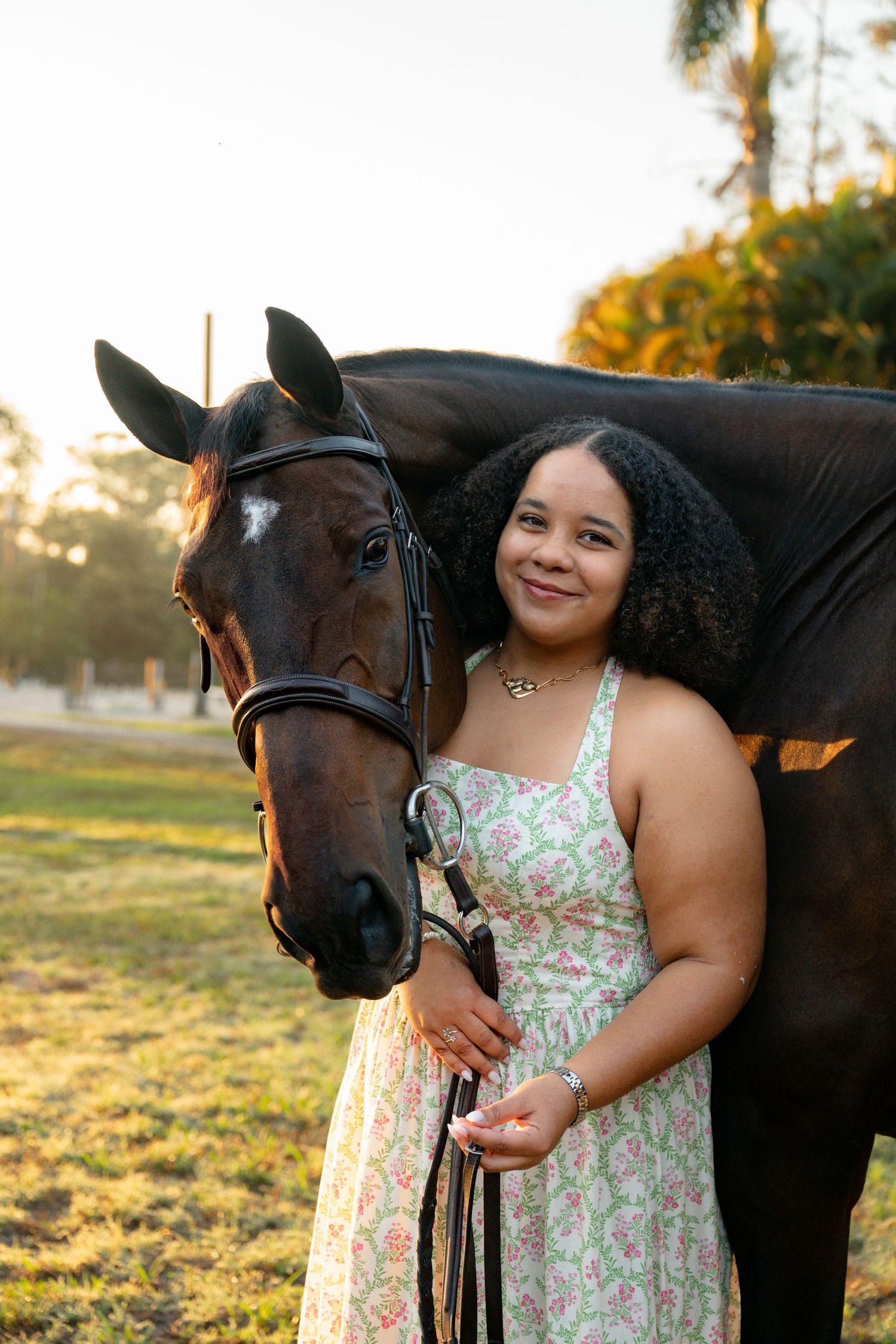 Smiling young woman in floral dress poses with a dark bay horse in a sunny outdoor paddock.