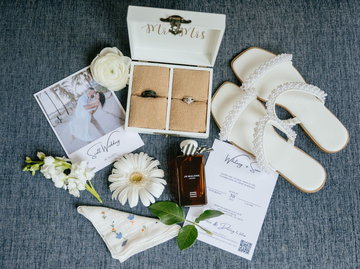 Flat lay of bridal accessories including rings, sandals, perfume, jewelry, and floral accents.