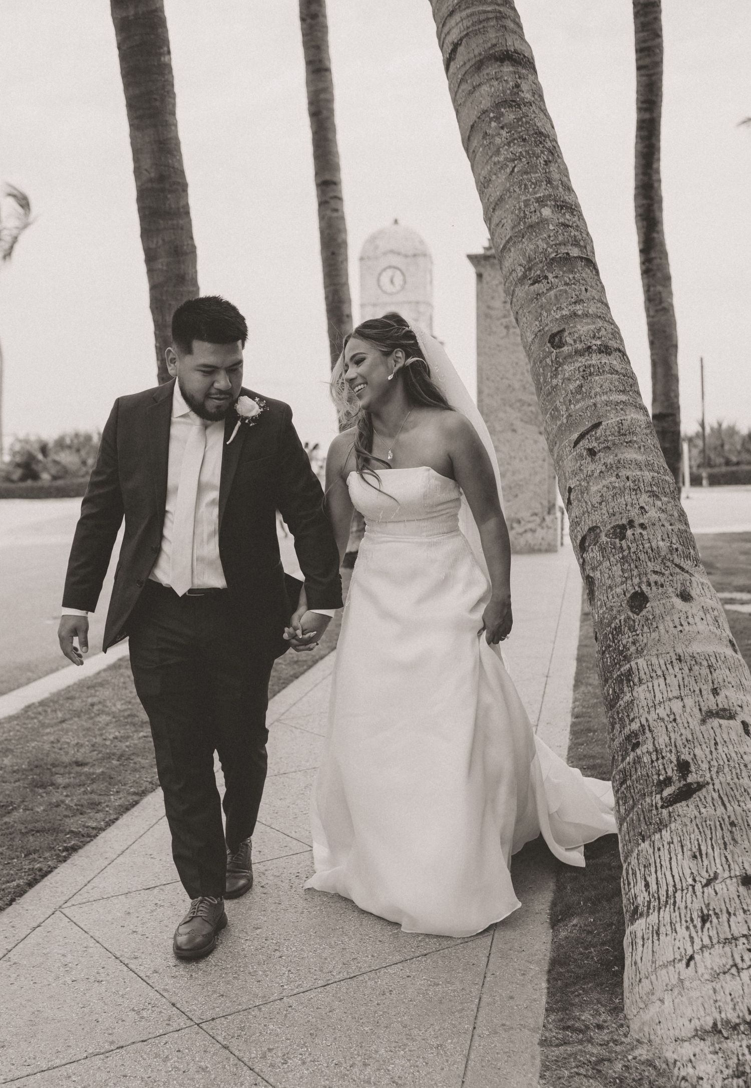 Black and white photo of bride and groom walking hand in hand past palm trees near clock tower.