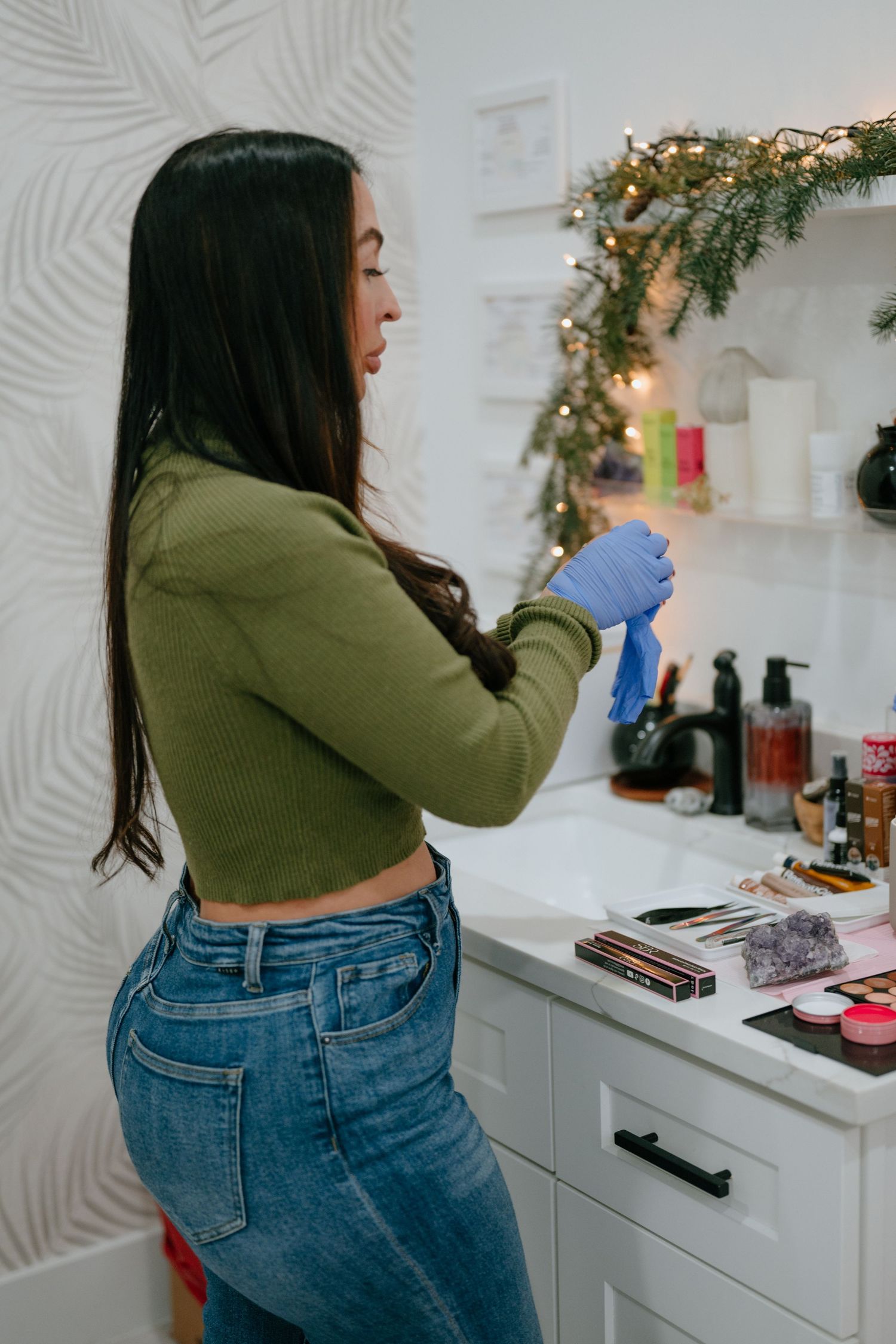 Female beauty technician in green crop top and jeans puts on blue latex gloves at a decorated makeup station.