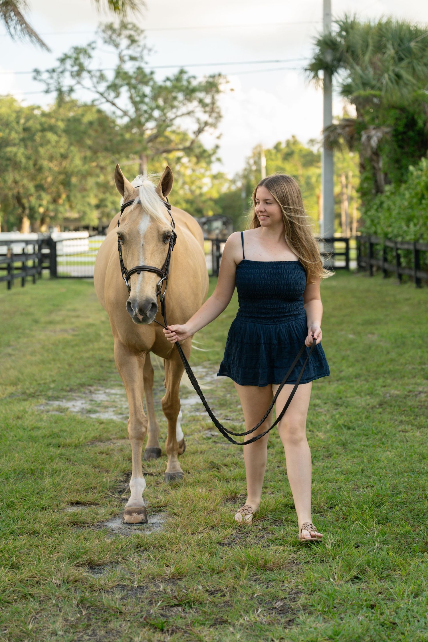 Young woman in navy dress walks palomino horse along grassy path beside black fence at horse farm.