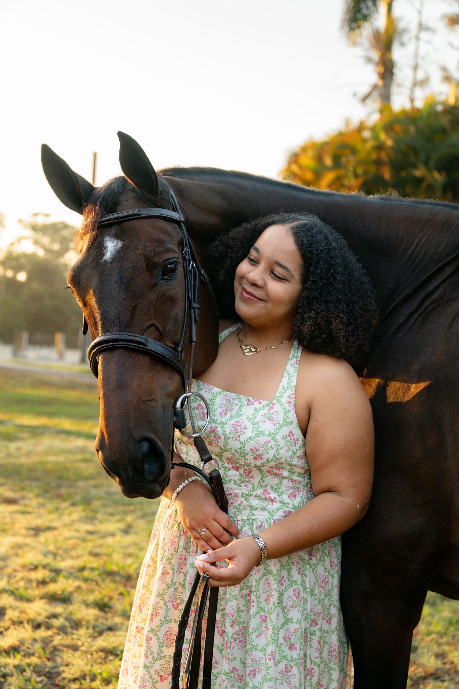 Smiling woman in floral dress stands closely beside a dark bay horse in warm golden sunlight.