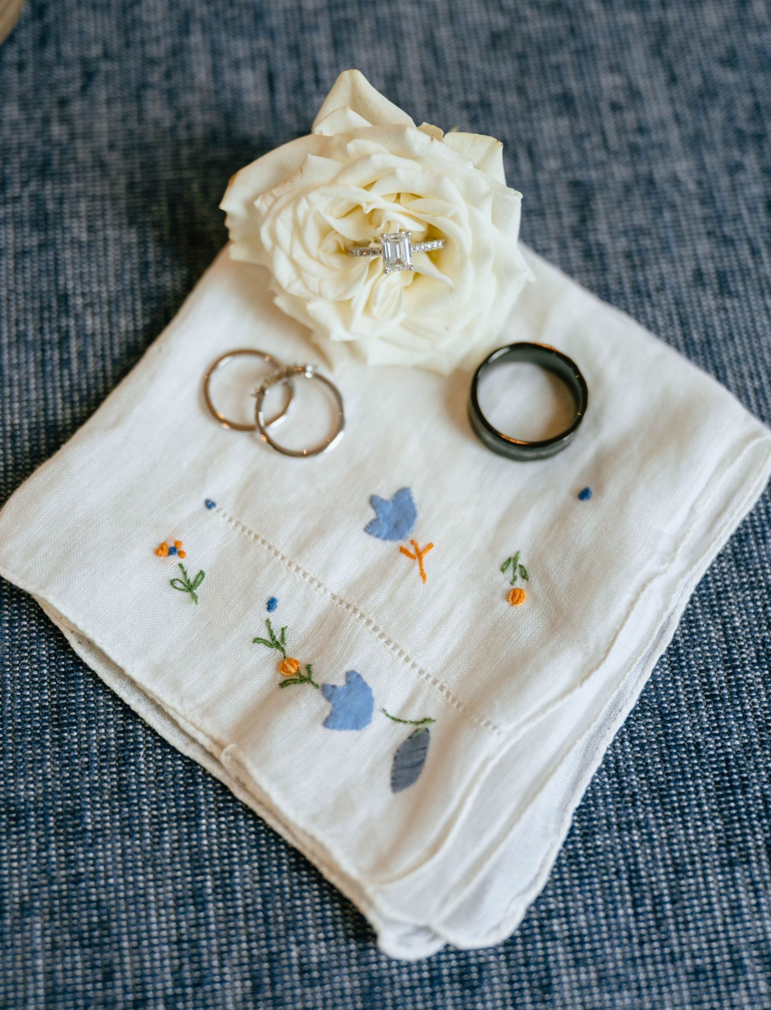 Wedding rings displayed on embroidered floral handkerchief with white rose accent on blue fabric.
