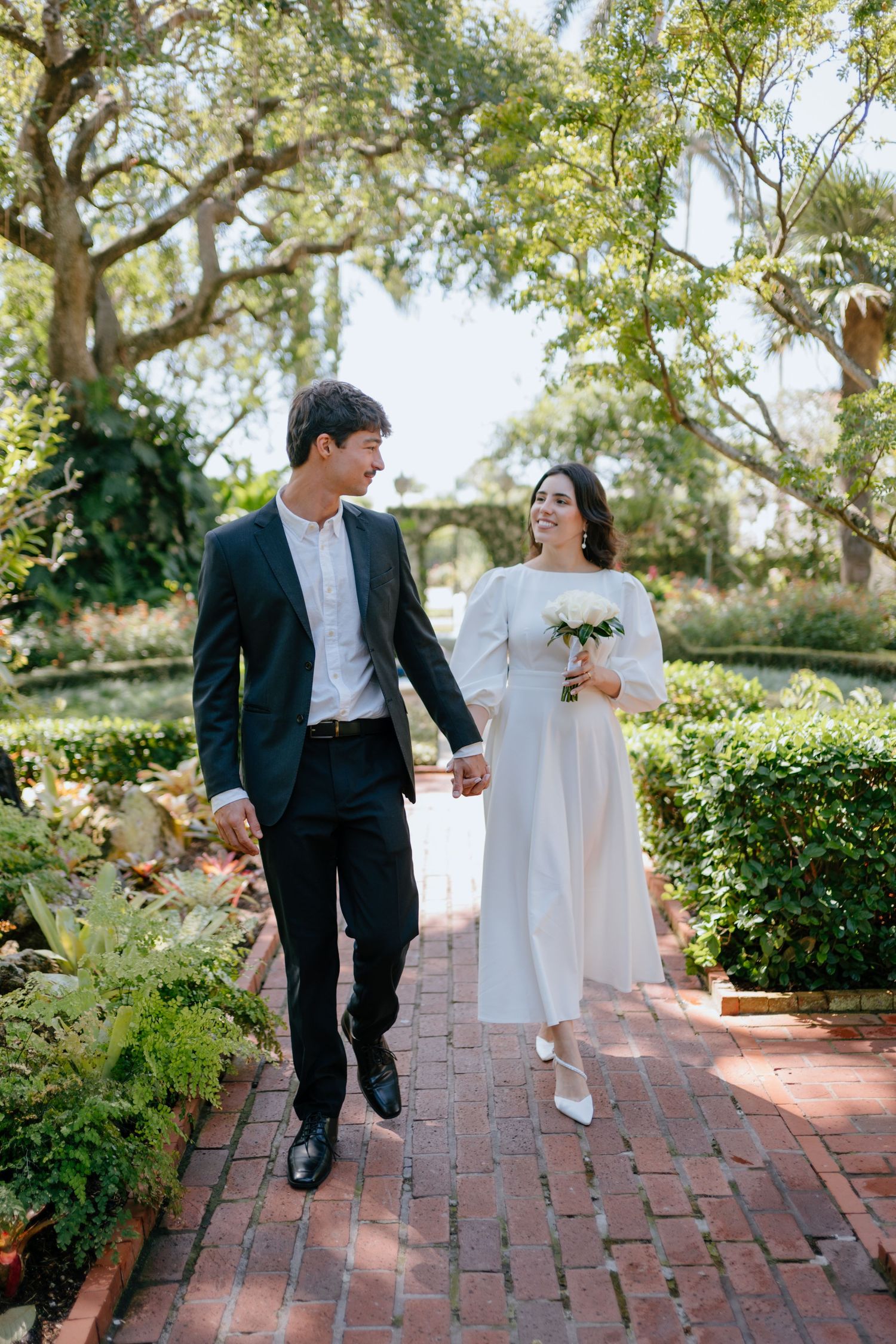 Smiling newlyweds walk hand in hand along brick garden path, bride holding white bouquet in lush setting.