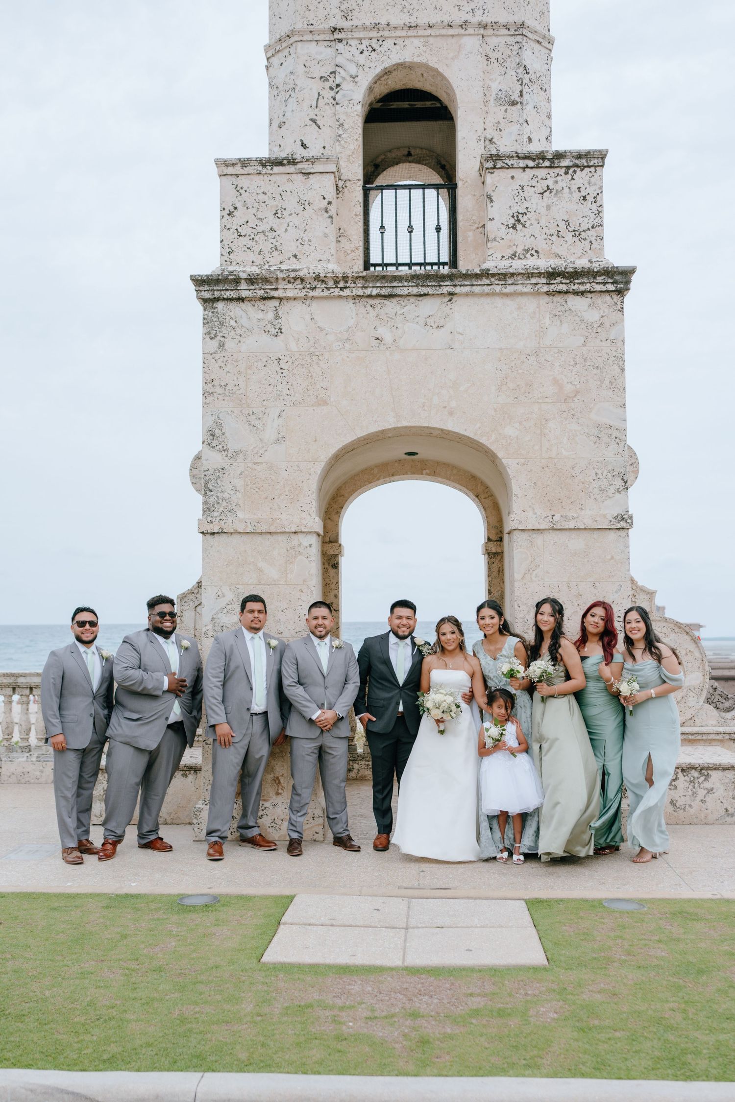 Wedding party poses before historic stone tower with ocean backdrop on couple's special day.