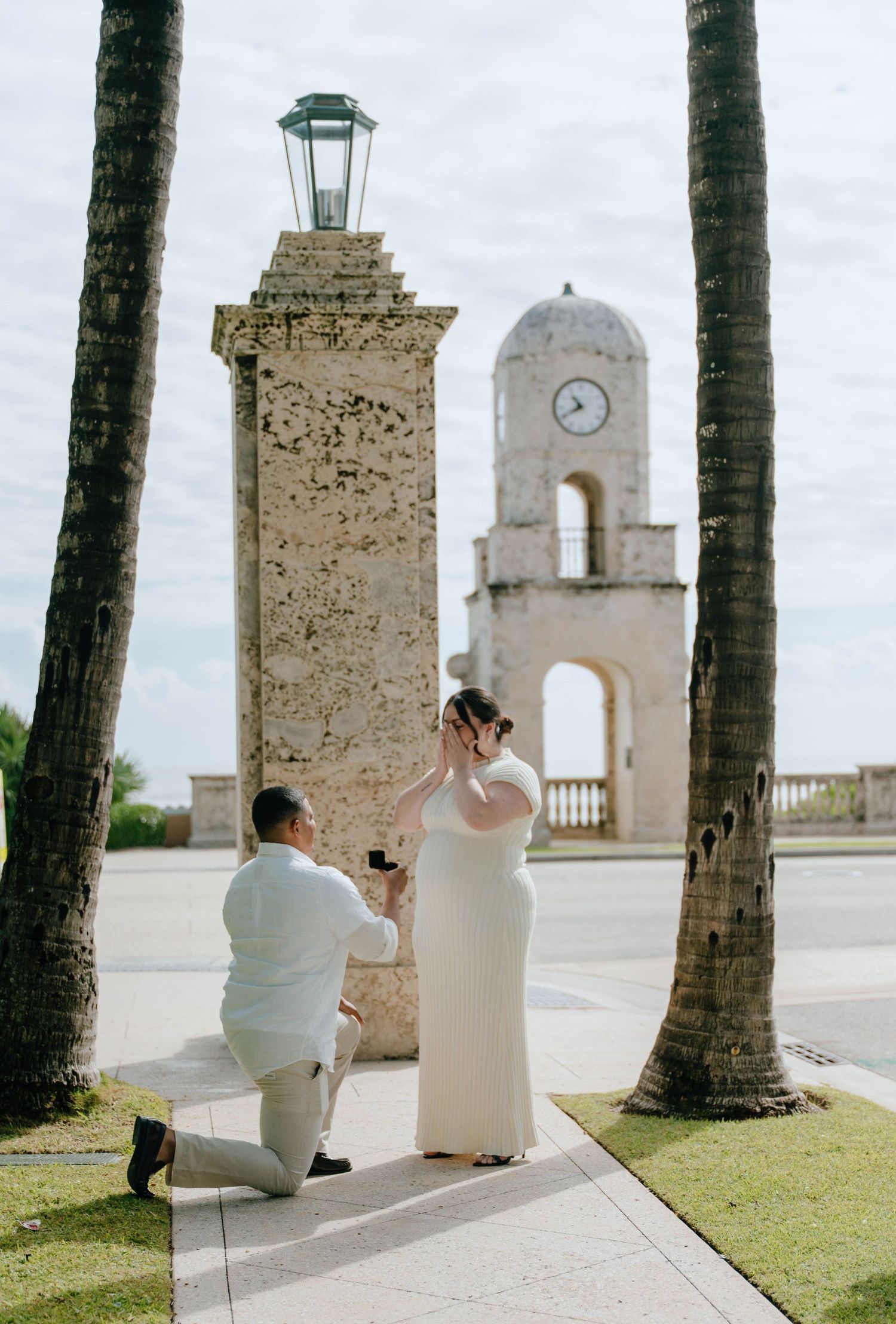 Man proposing on one knee to woman in white dress near a historic clock tower with palm trees.