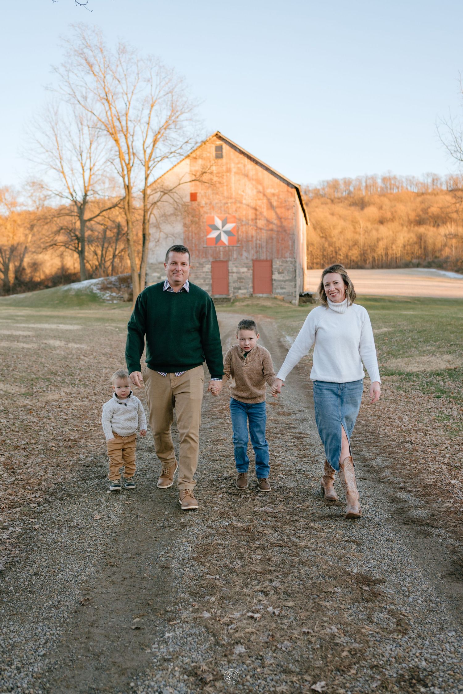 Smiling family of four walking hand-in-hand on a gravel path with a rustic red barn in background.