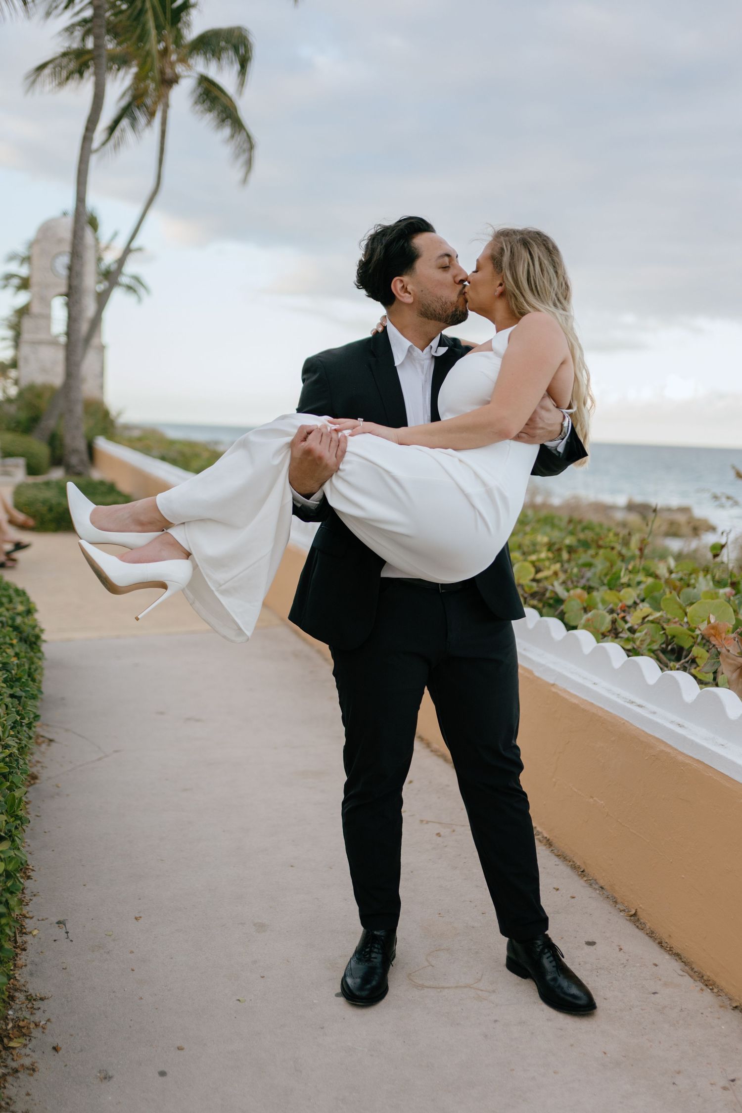 Groom in black suit lifting and kissing bride in white dress on a seaside pathway.