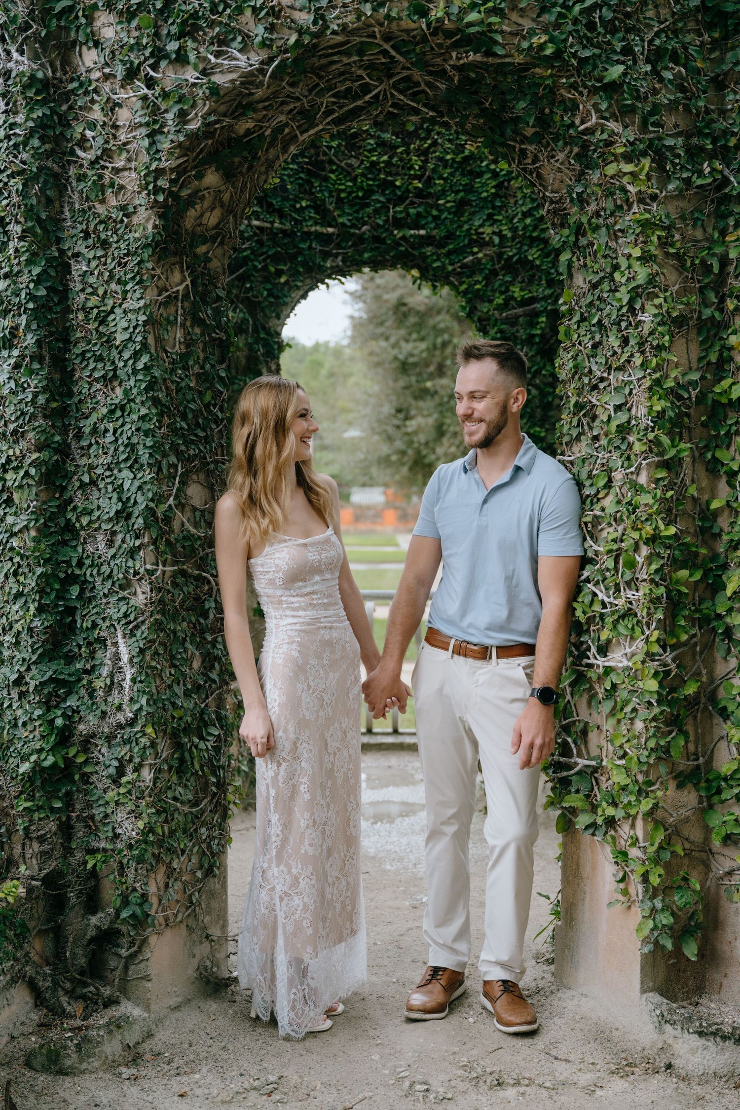 Smiling couple holding hands under an ivy-covered stone arch in a garden.