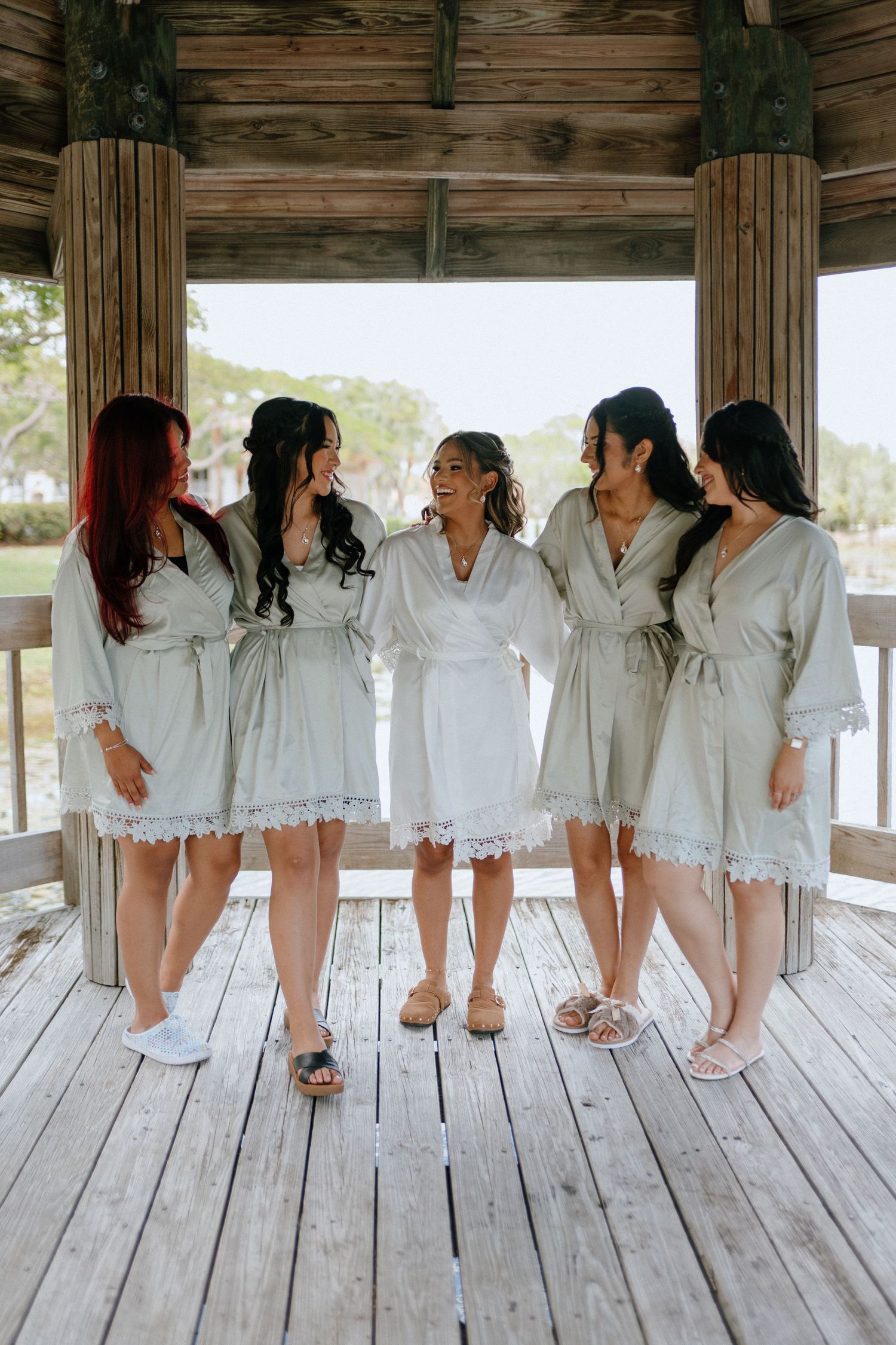 Bride and four bridesmaids laugh together wearing matching sage robes on a wooden deck.