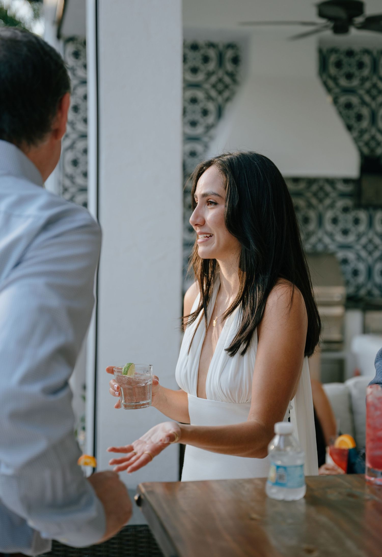 Bride in a white dress smiling and holding a cocktail while conversing at a bar during a wedding reception.