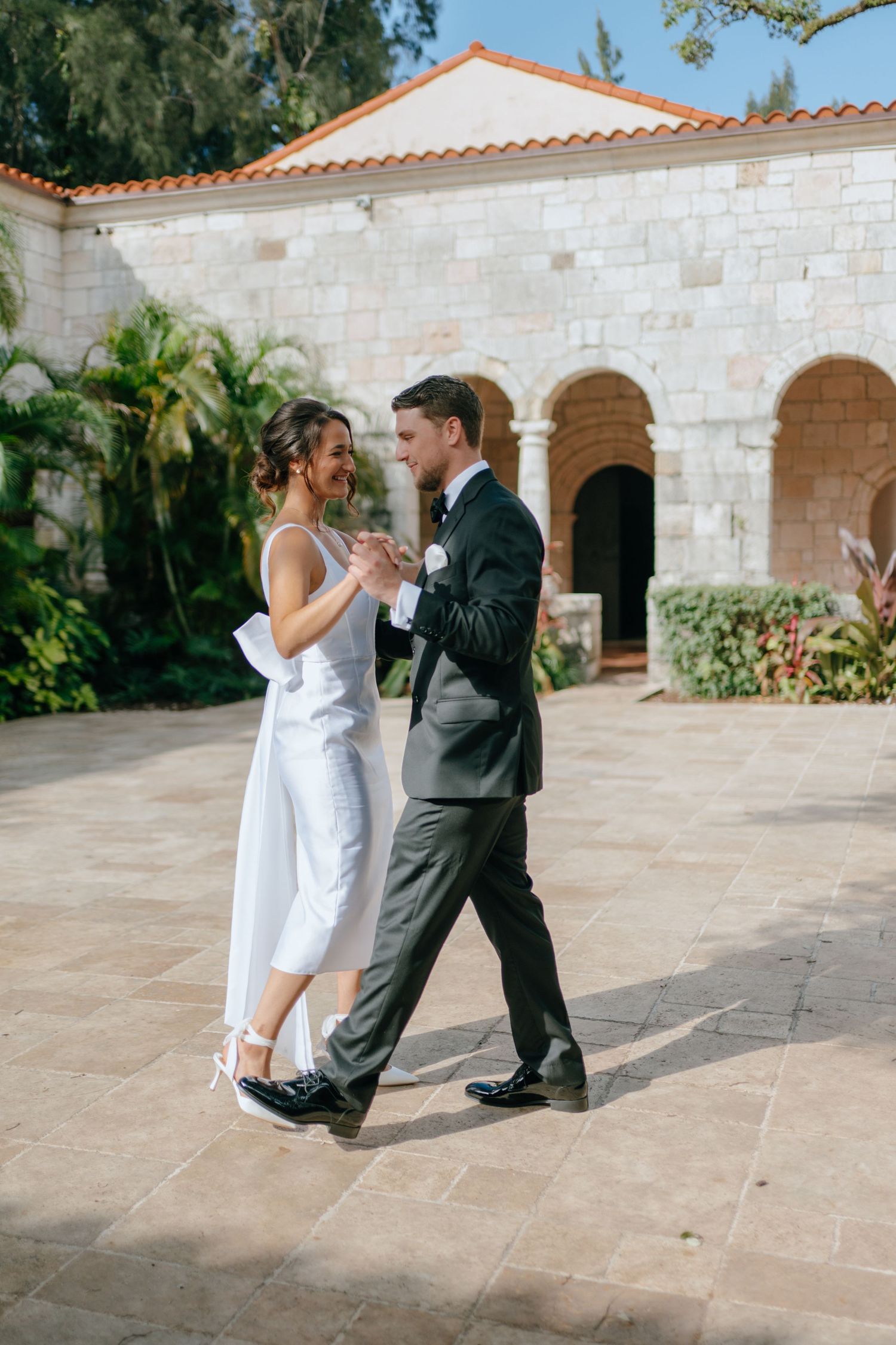 Bride in white jumpsuit and groom in dark suit dancing in a stone courtyard.
