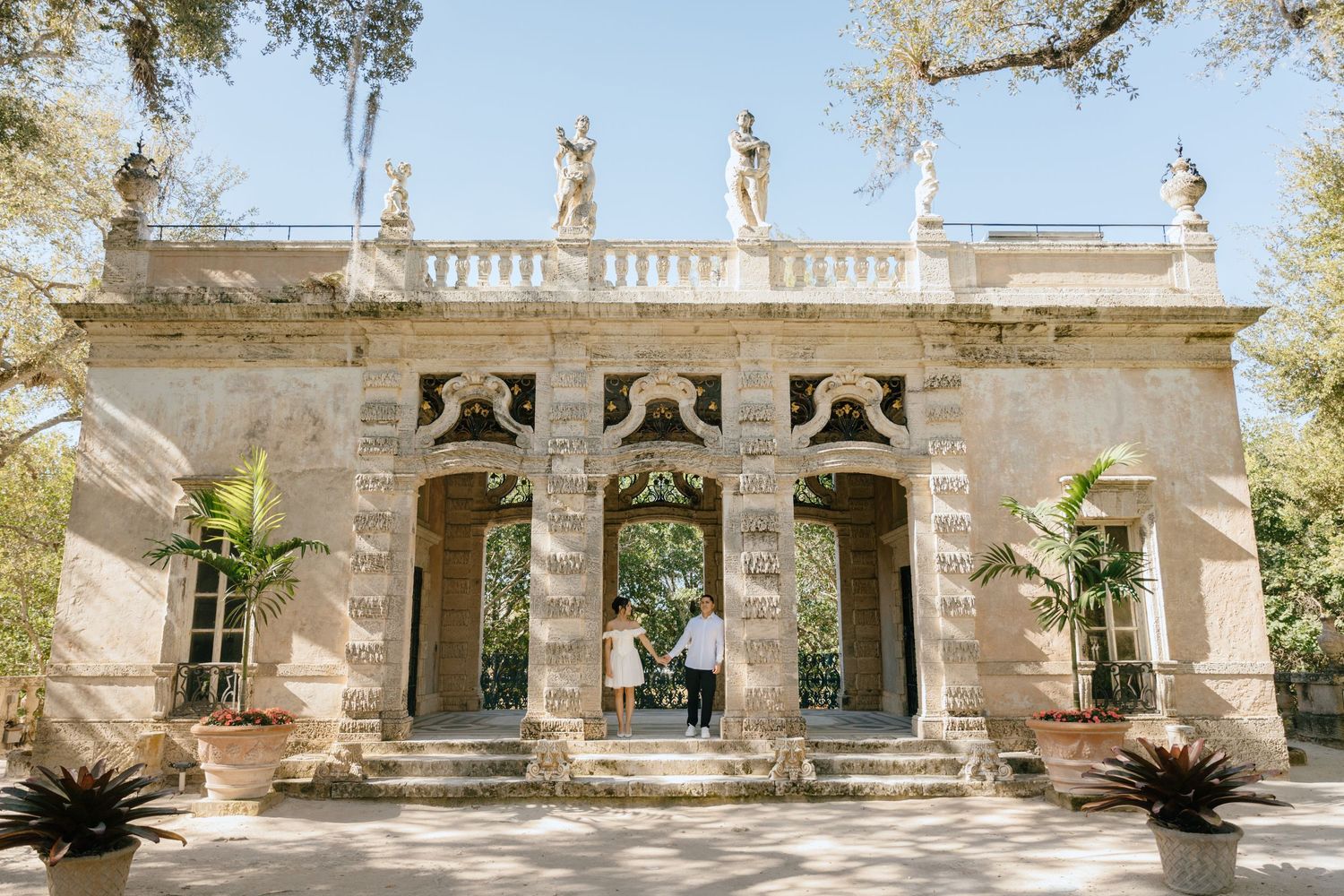 Couple stands together beneath ornate stone archways of a classical Mediterranean-style villa.