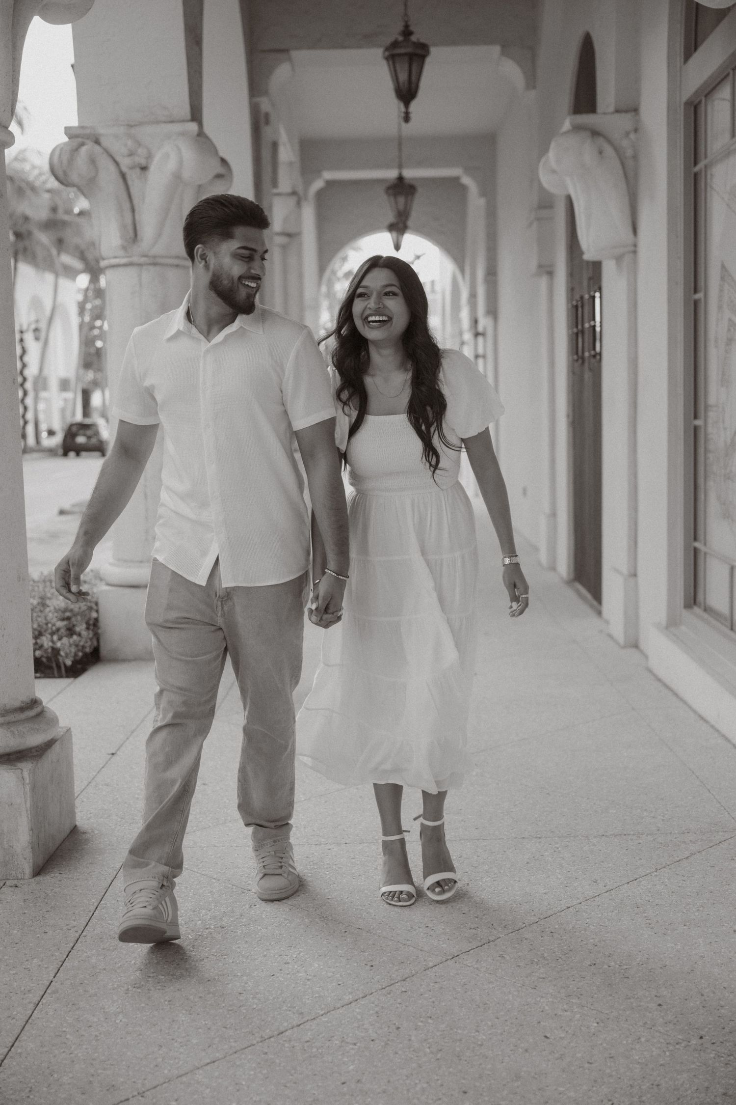 Happy couple in white outfits walking hand in hand through an elegant columned corridor.