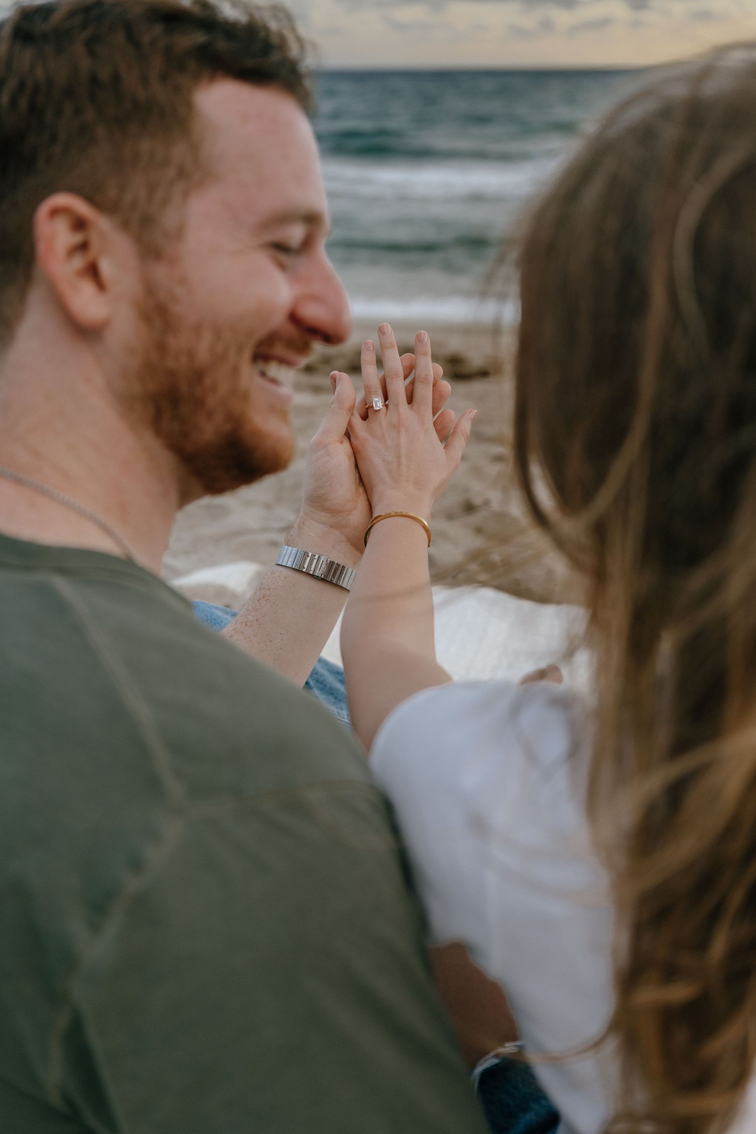 Smiling bearded man admiring engagement ring on womans hand at beach after proposal.