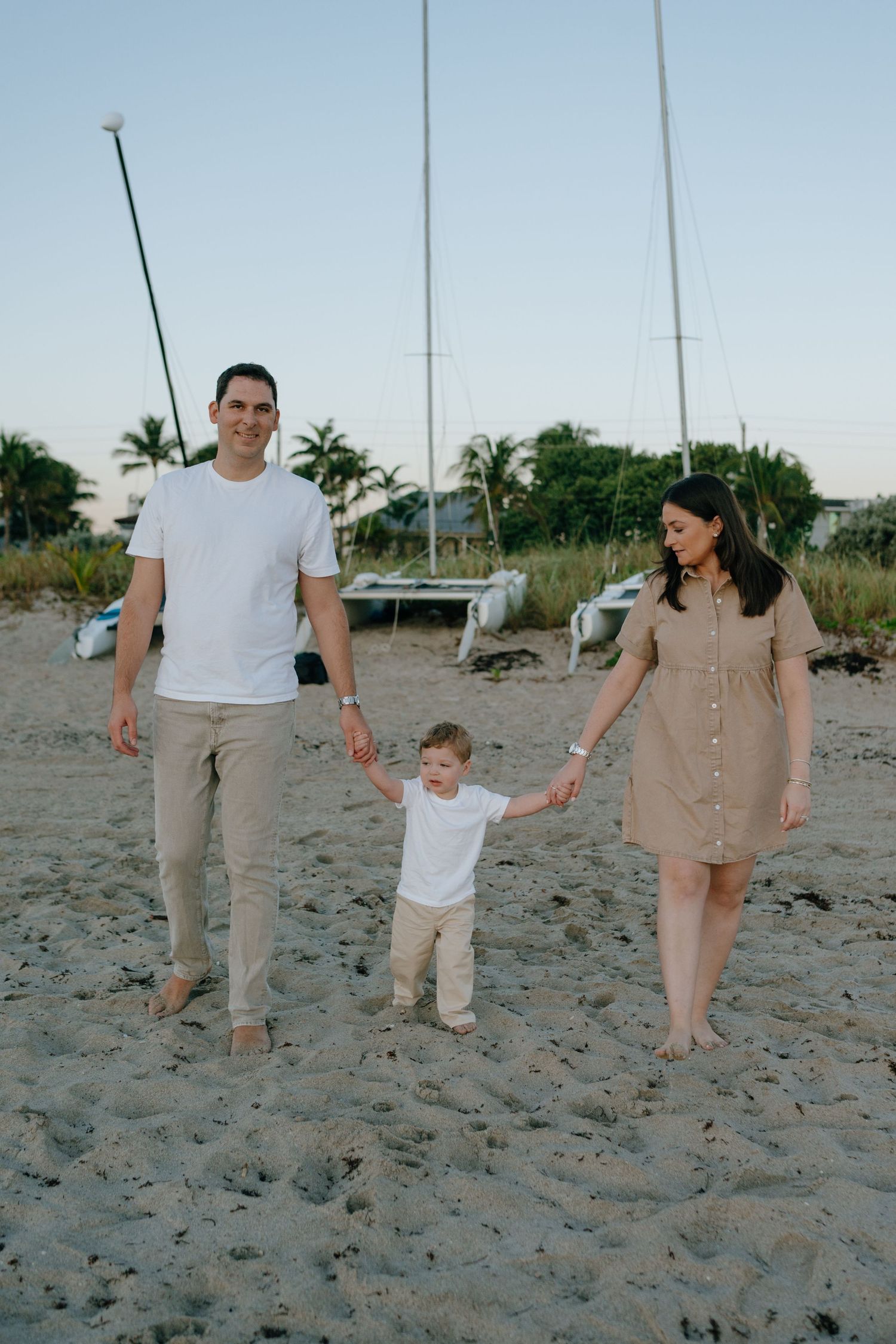 Young family walking hand in hand with their toddler on a sandy beach with sailboats in the background.