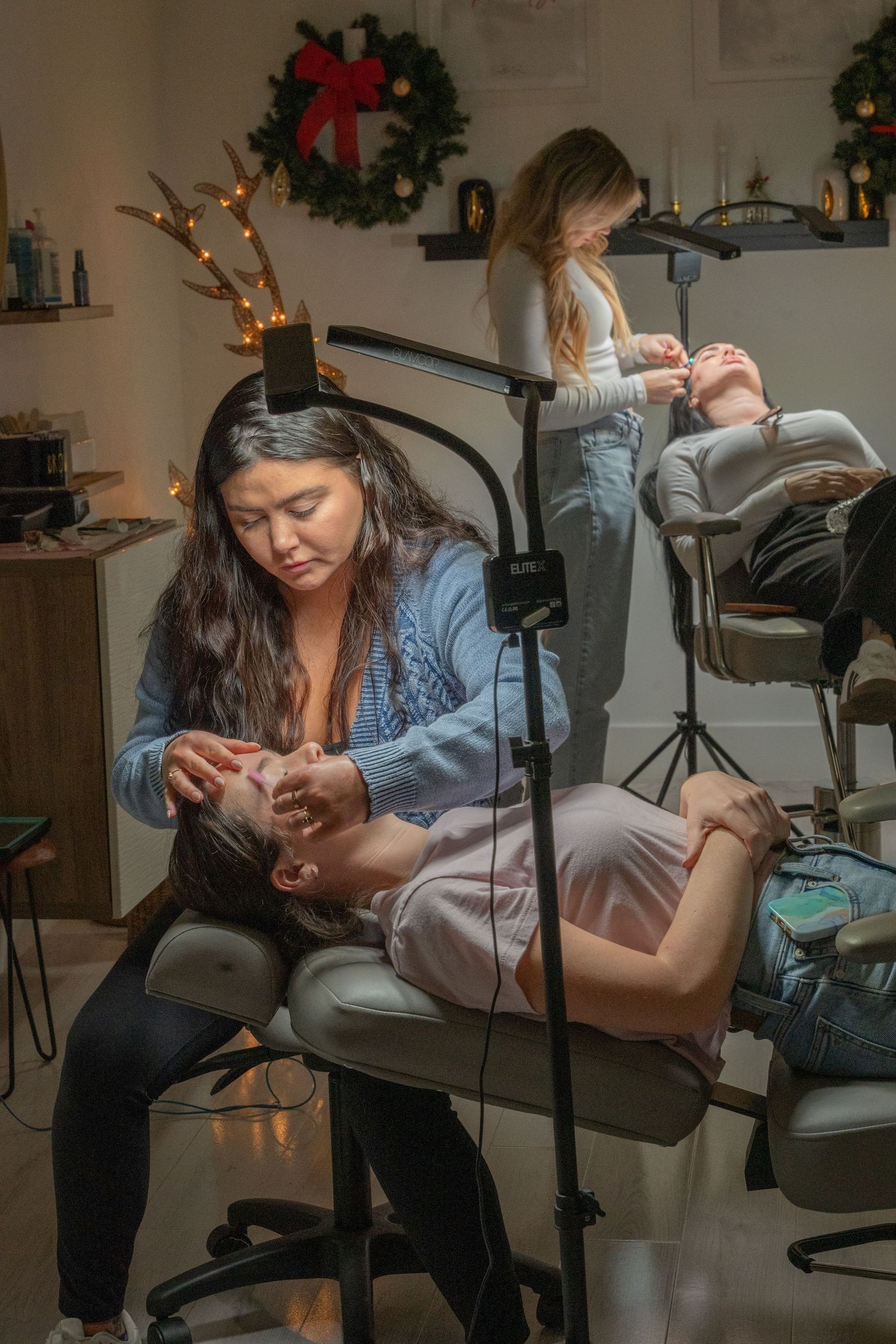 Two estheticians performing eyebrow treatments on clients in a festively decorated beauty salon.