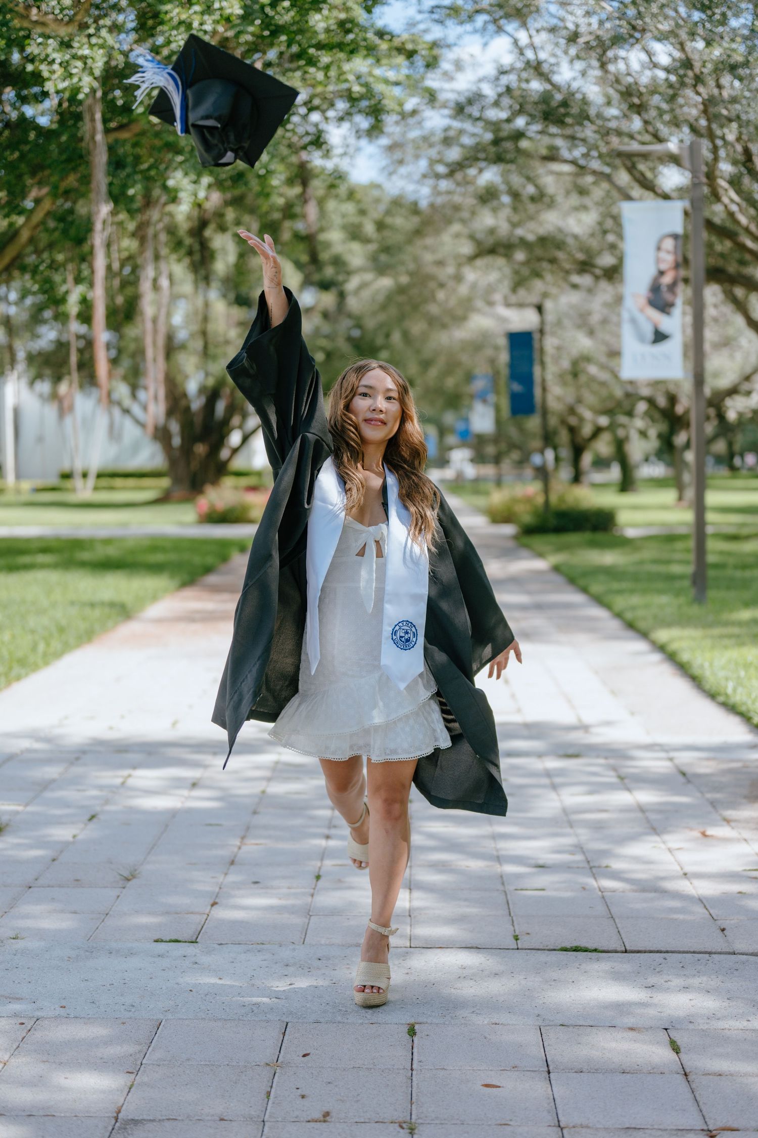 Excited graduate tossing black cap in the air while wearing gown on a sunny campus walkway.