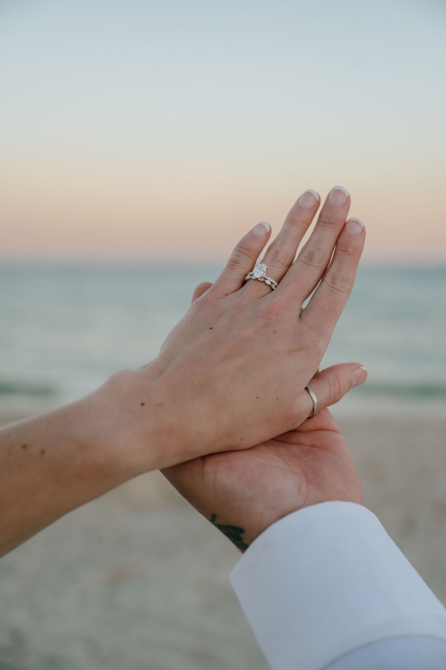 Couple holding hands on beach at sunset showing engagement and wedding rings.