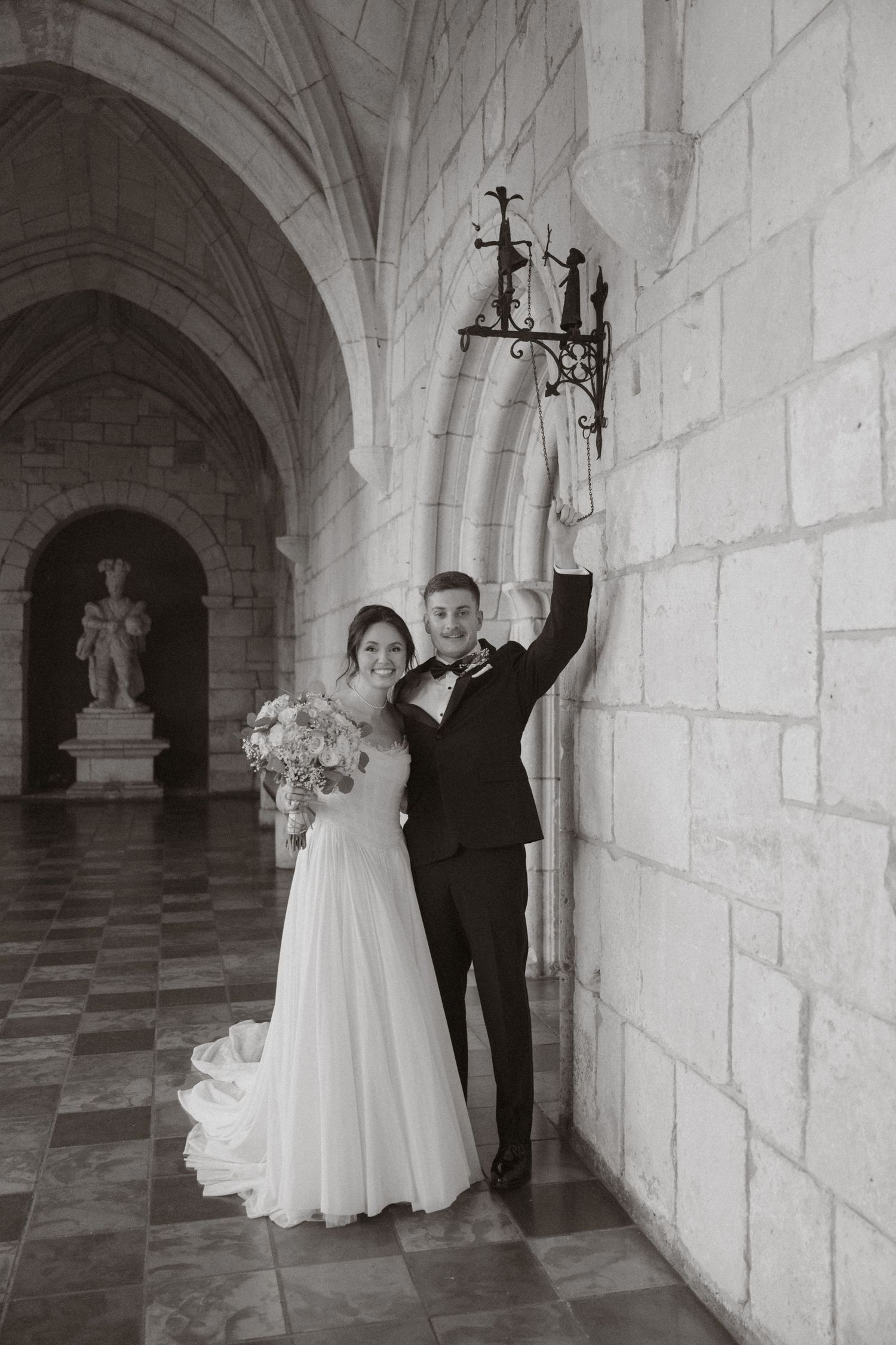 Black and white photo of smiling bride and groom posing in ornate stone cathedral corridor.