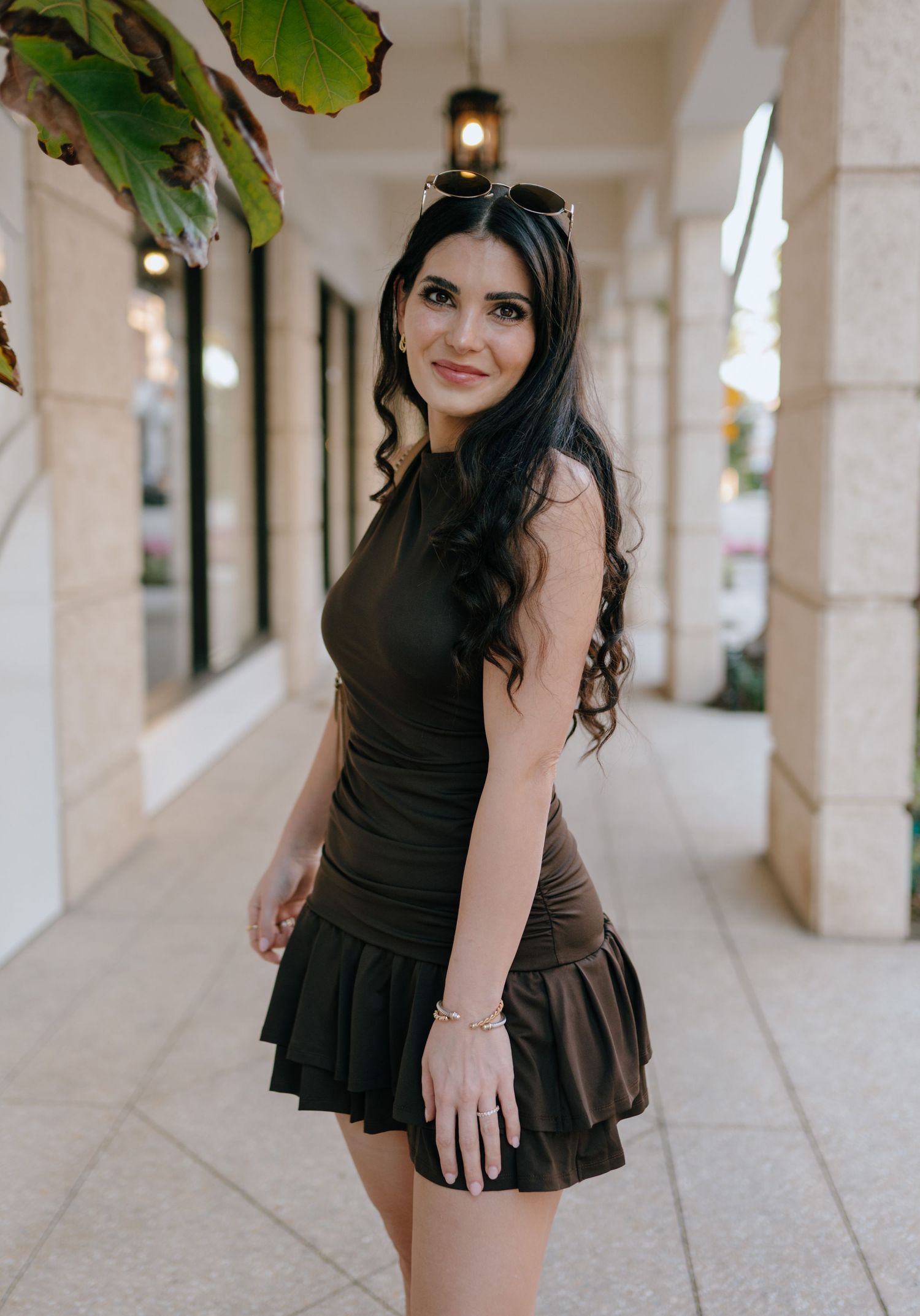 A smiling brunette woman in a dark mini dress posing in a sunlit outdoor walkway.