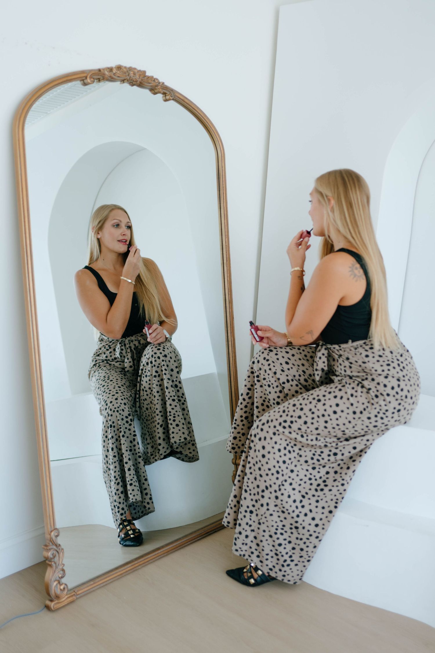 Blonde woman in black top and leopard pants applying lipstick at ornate floor mirror.
