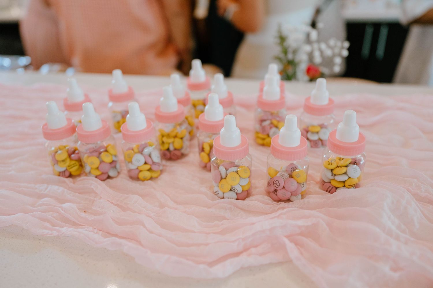 Pink baby bottle favors filled with colorful candies displayed on pink fabric at a baby shower.