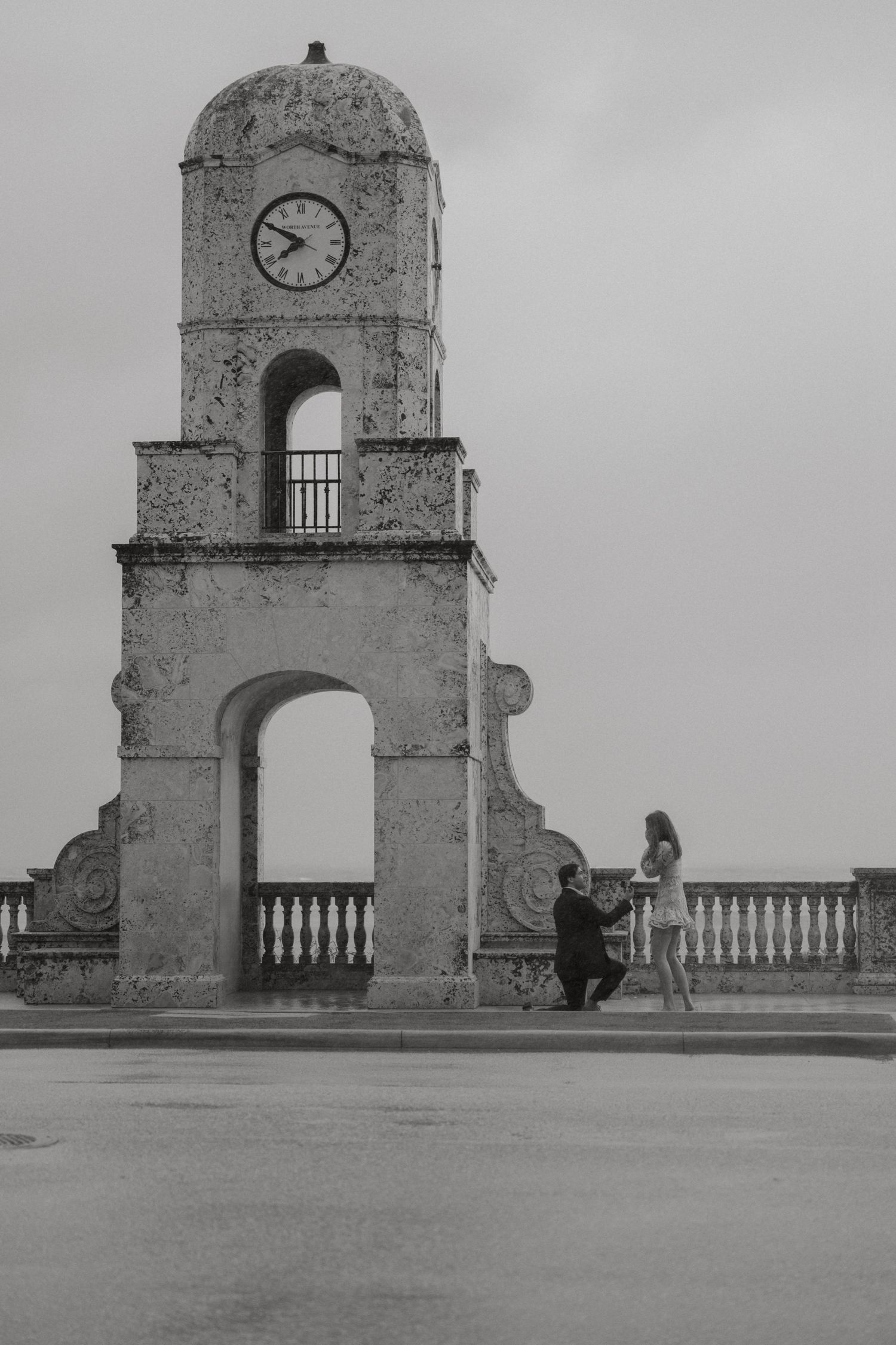 Man proposing to his partner on one knee near a historic clock tower on a cloudy day.