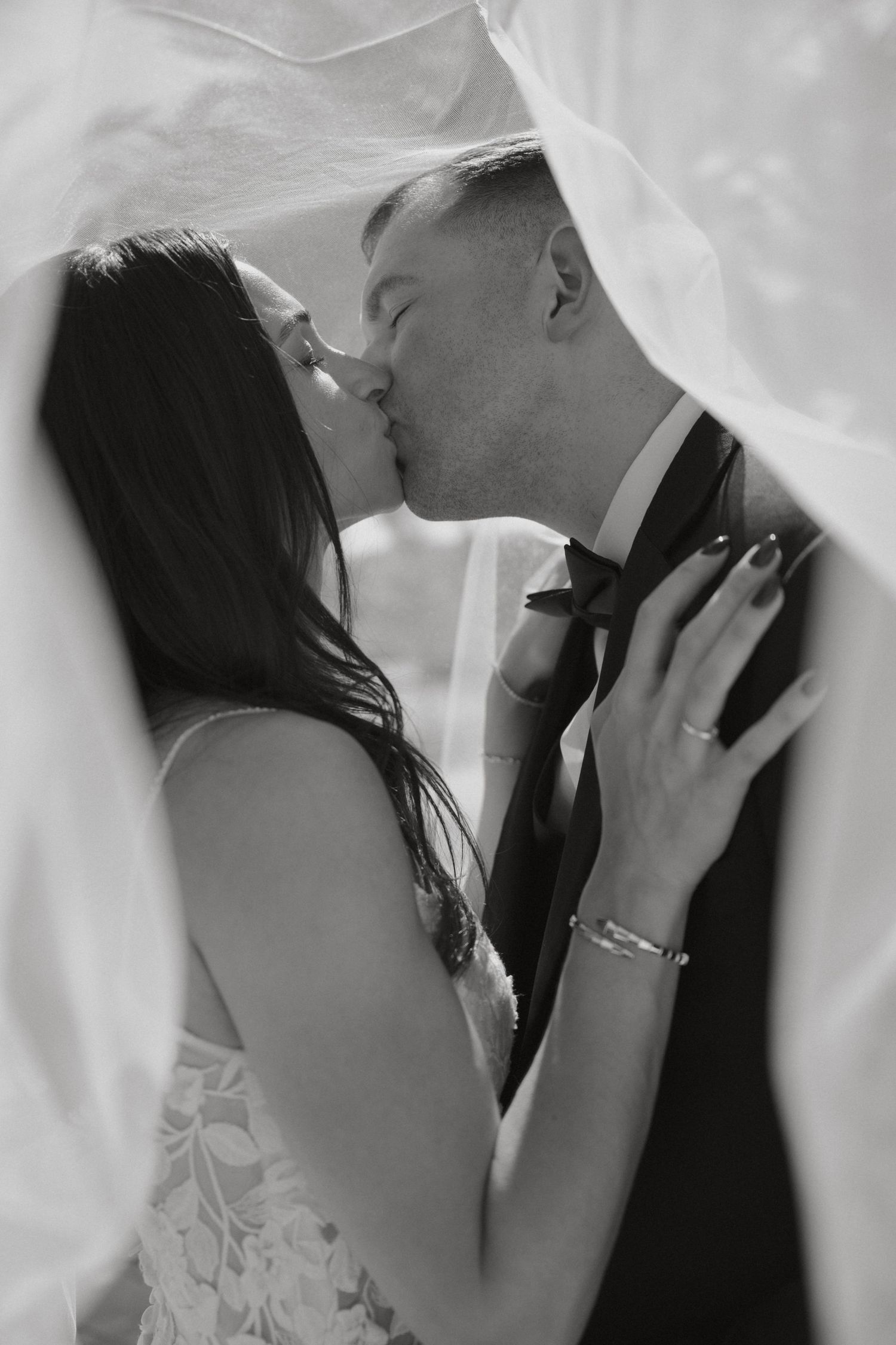 Black and white romantic photo of bride and groom kissing under a flowing white veil.