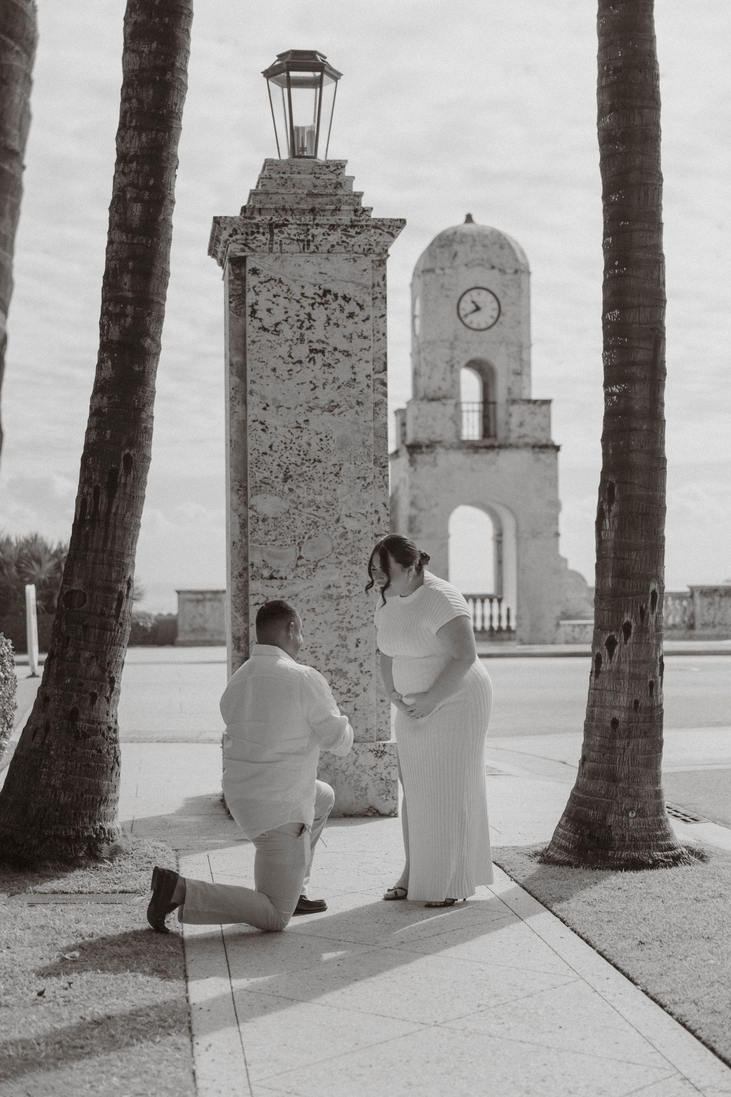 Black and white photo of marriage proposal near historic clock tower framed by palm trees.