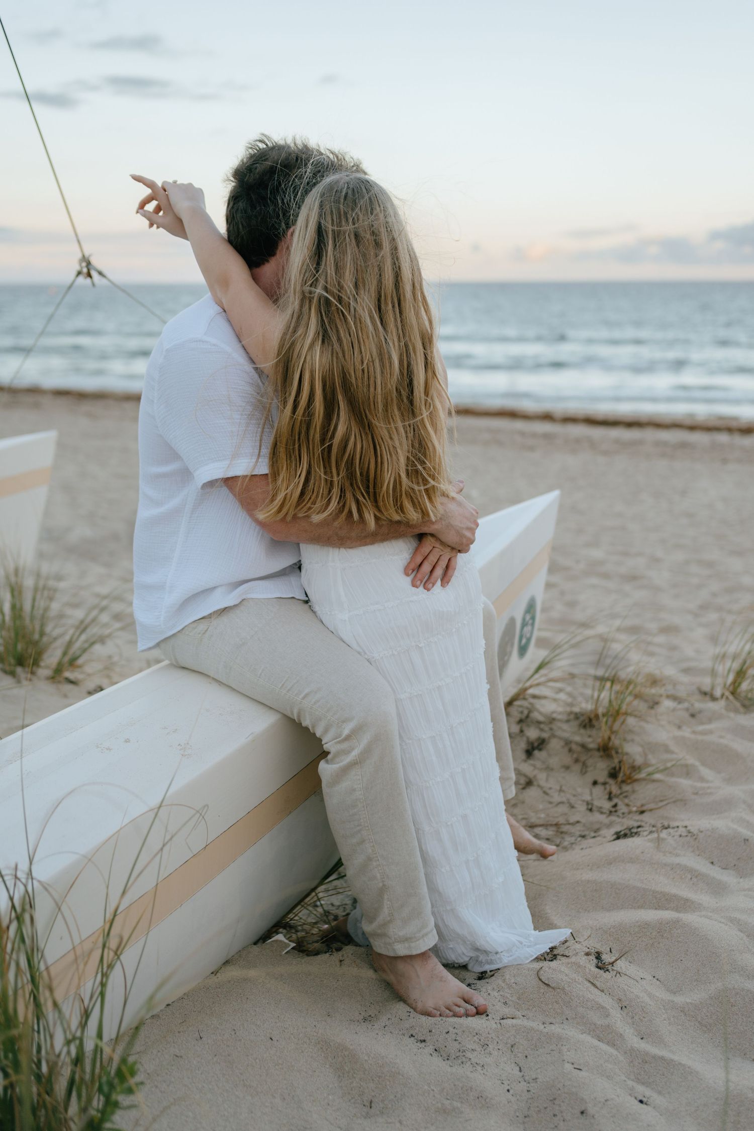 Couple sitting together on a beach bench at sunset, embracing and pointing toward the ocean horizon.