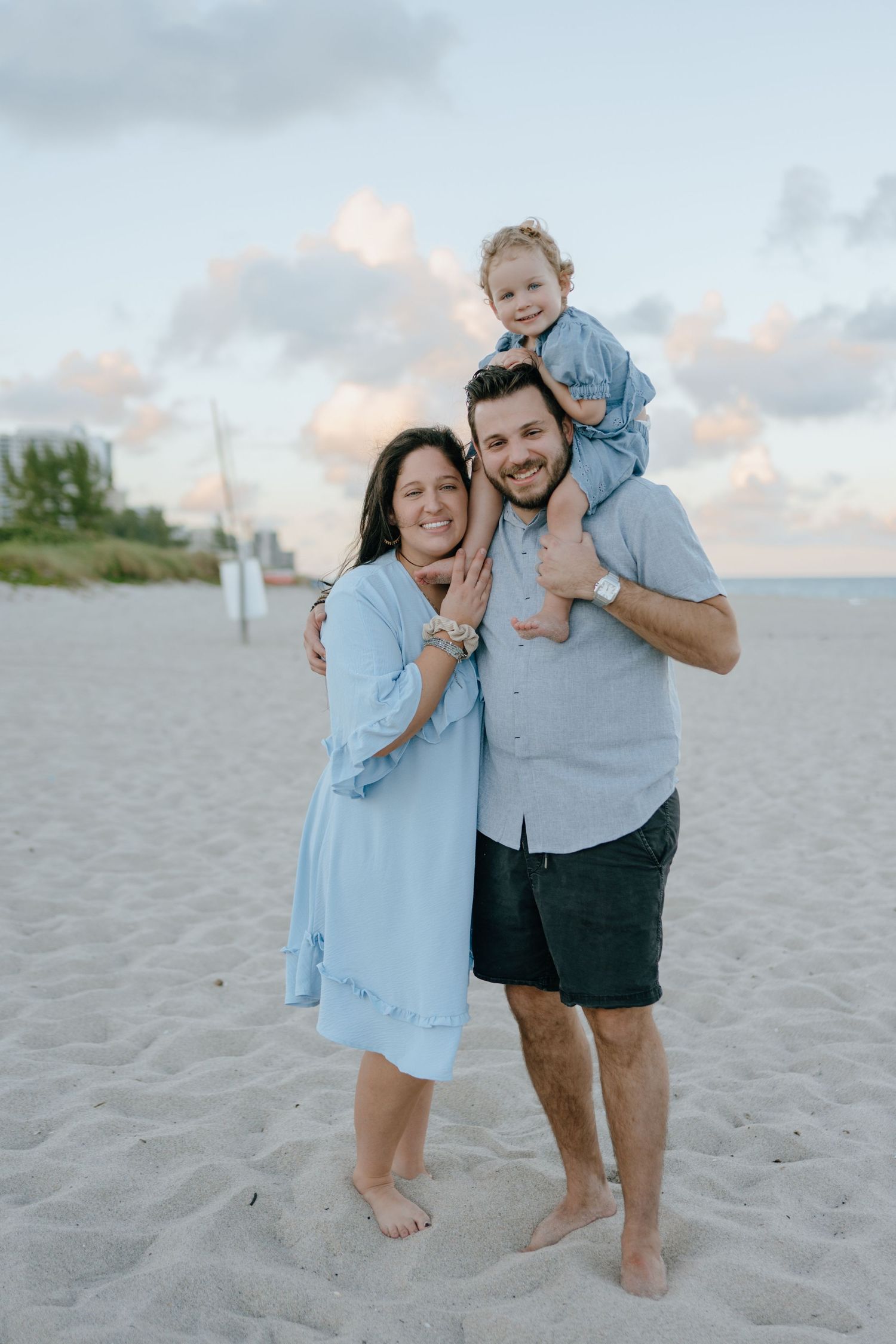 Smiling family of three posing on the beach at dusk, with toddler girl sitting on fathers shoulders.