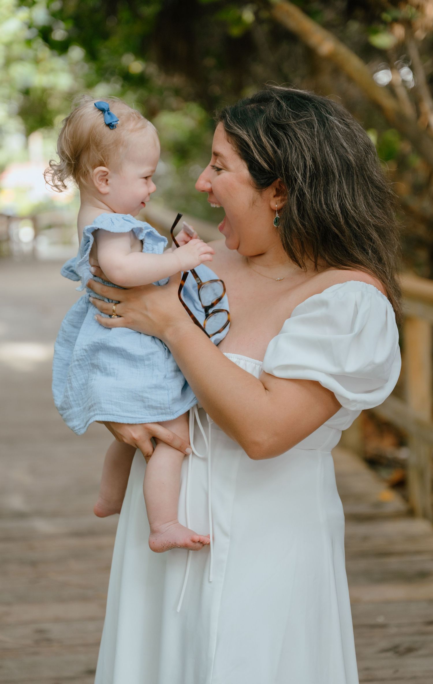 A laughing woman in a white dress holds a smiling baby girl on a sunny wooden boardwalk.