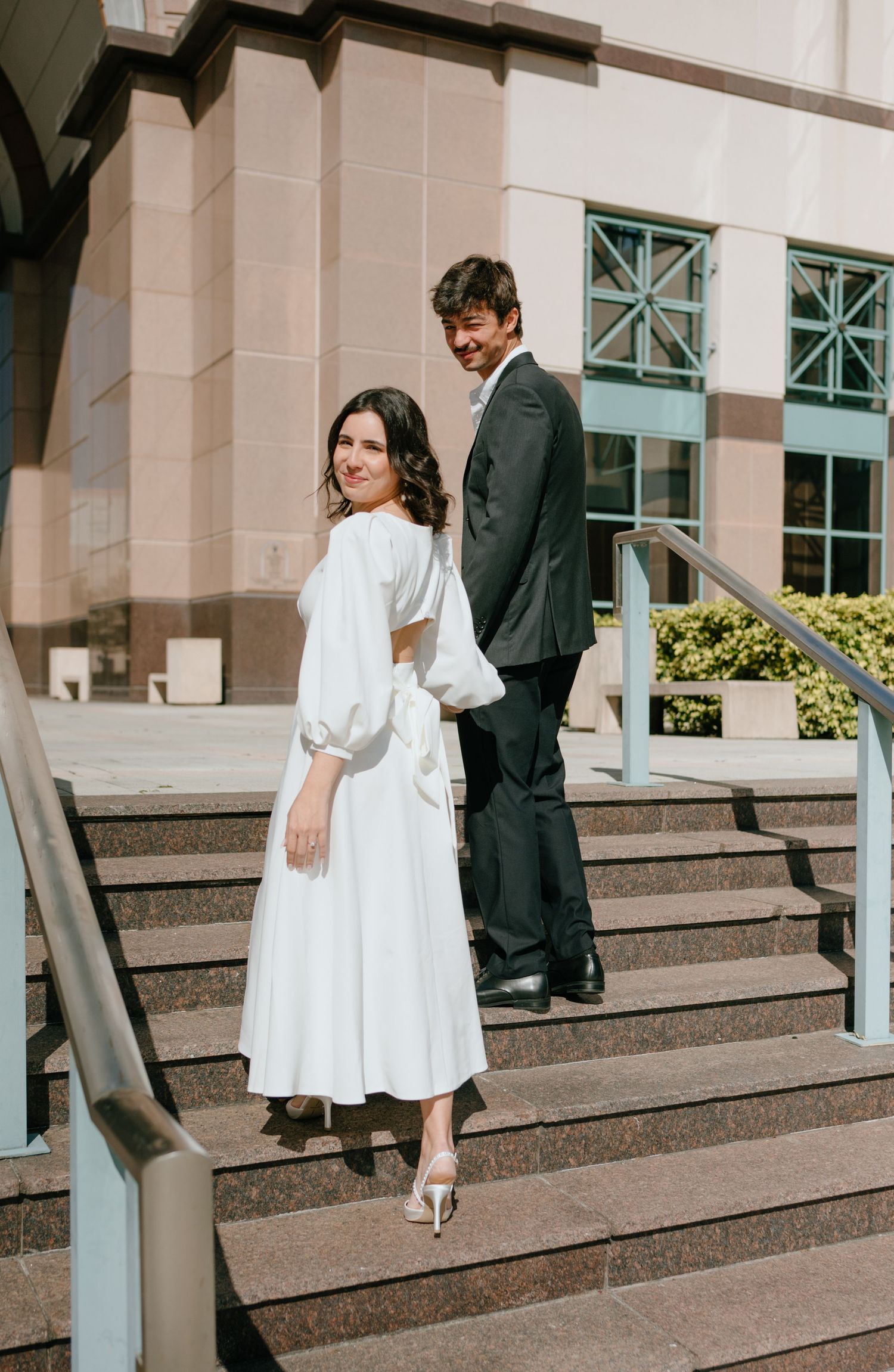 Elegant bride in white midi dress and groom in dark suit pose on courthouse steps for wedding photos.