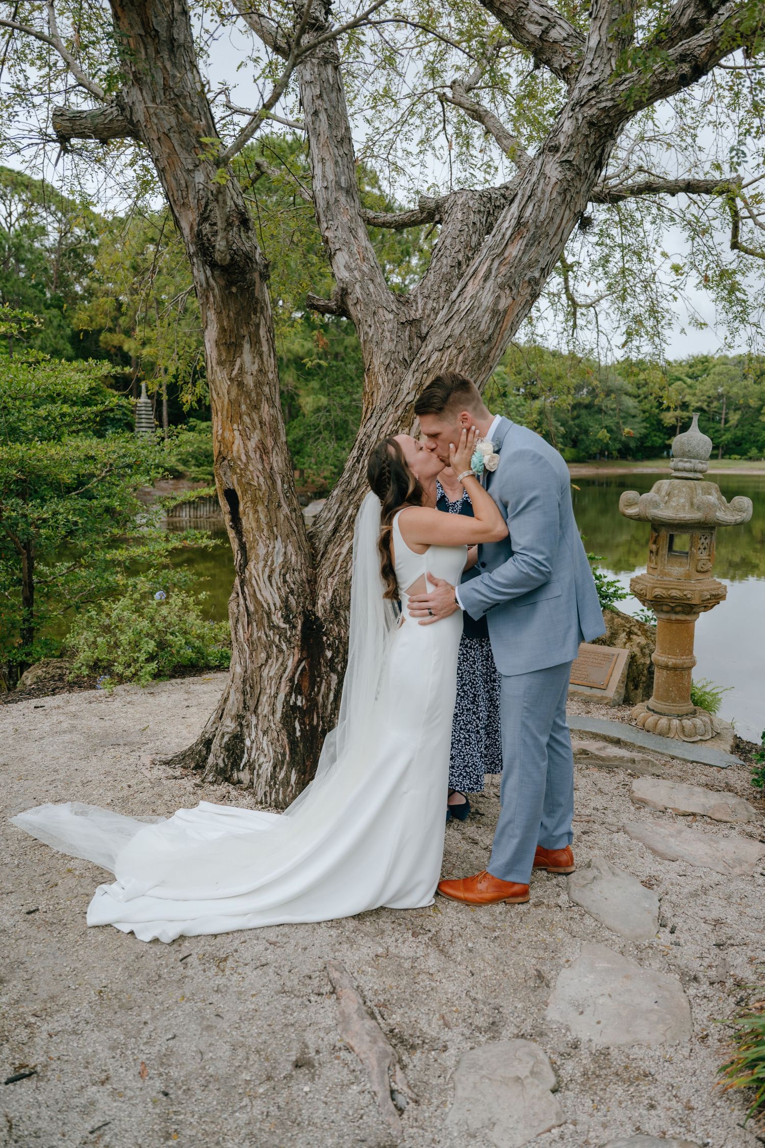 Newlyweds share an intimate embrace under a large tree by a tranquil pond in a garden setting.