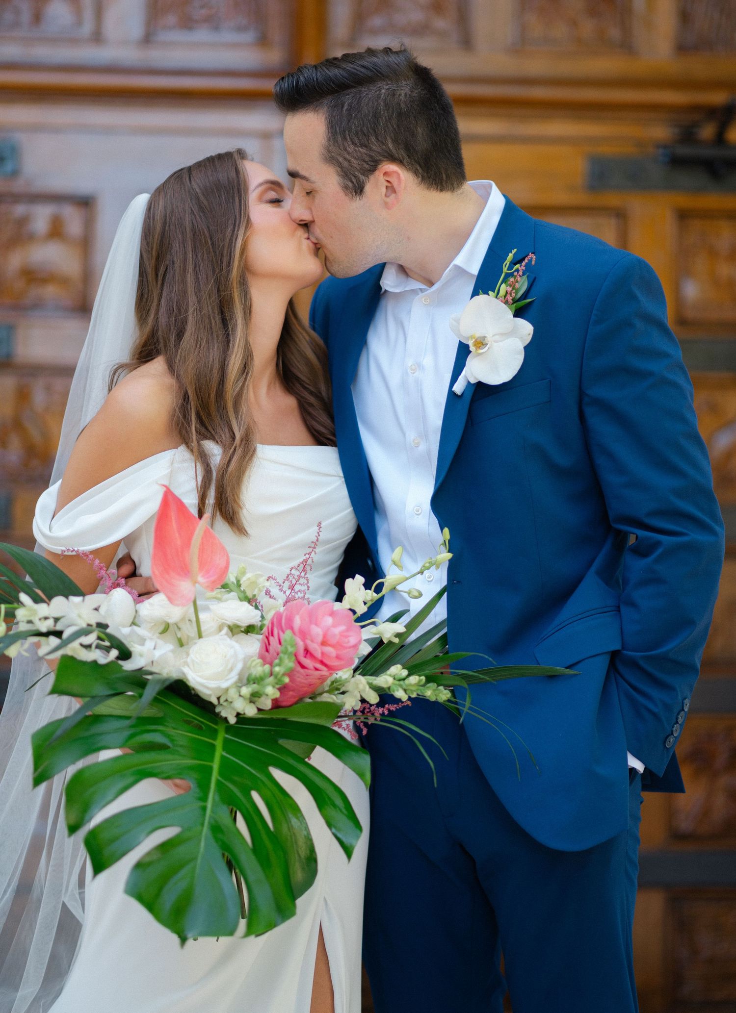 Newlyweds share a first kiss holding a tropical floral bouquet with pink and white flowers.