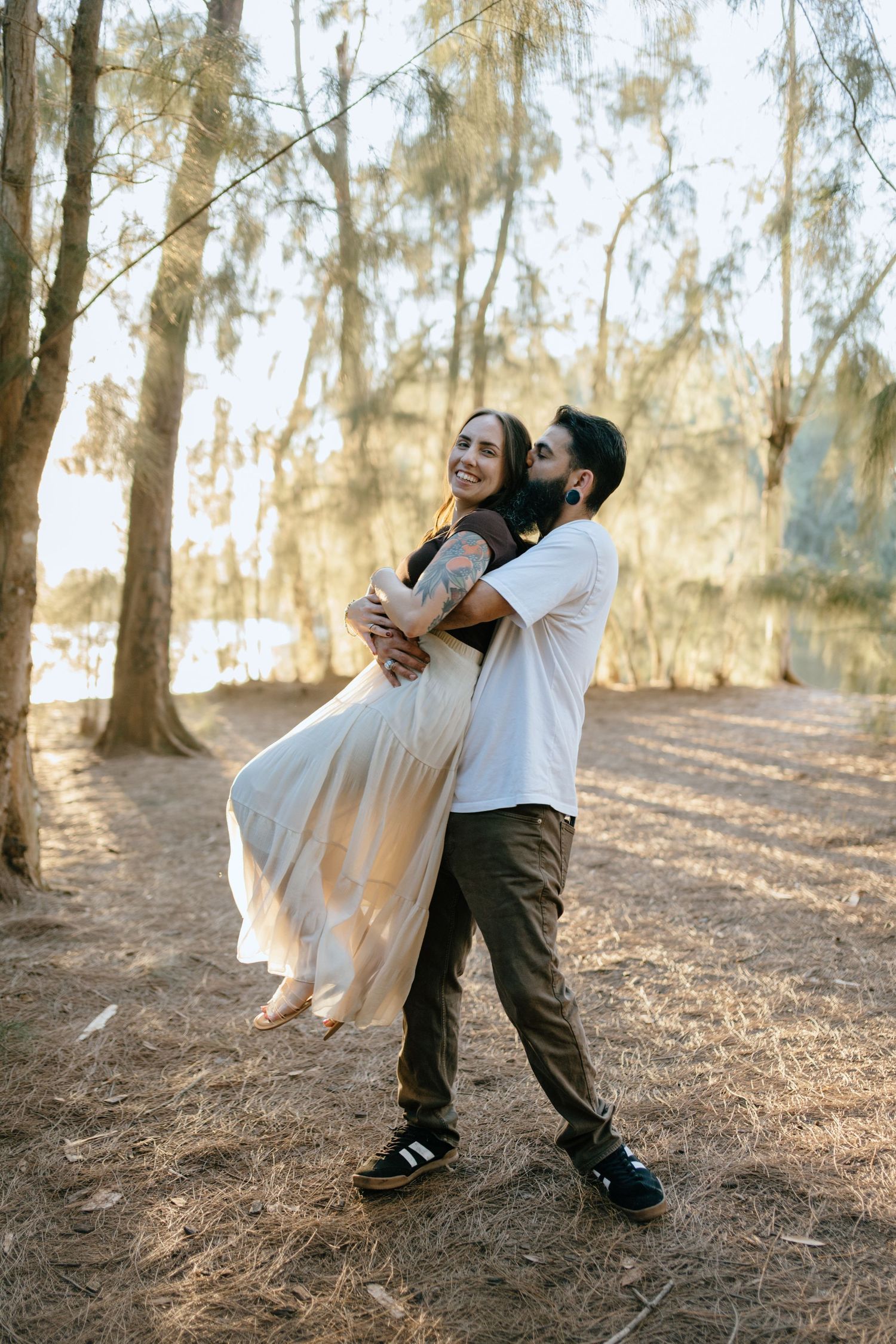 Man lifts smiling woman in flowing white skirt in a warm, sunlit forest setting.