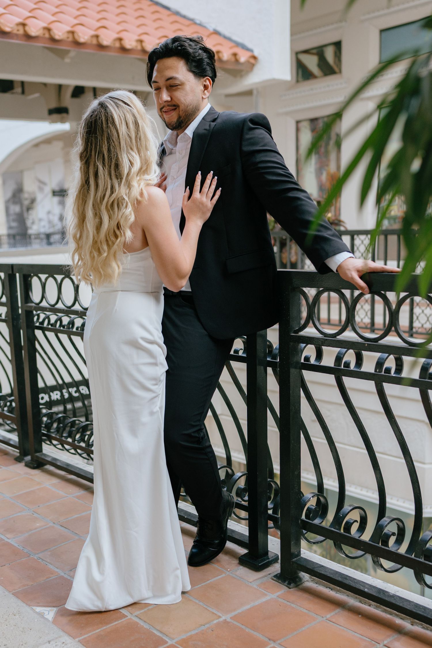 Bride in white gown and groom in black suit sharing a romantic moment on a balcony with iron railings.