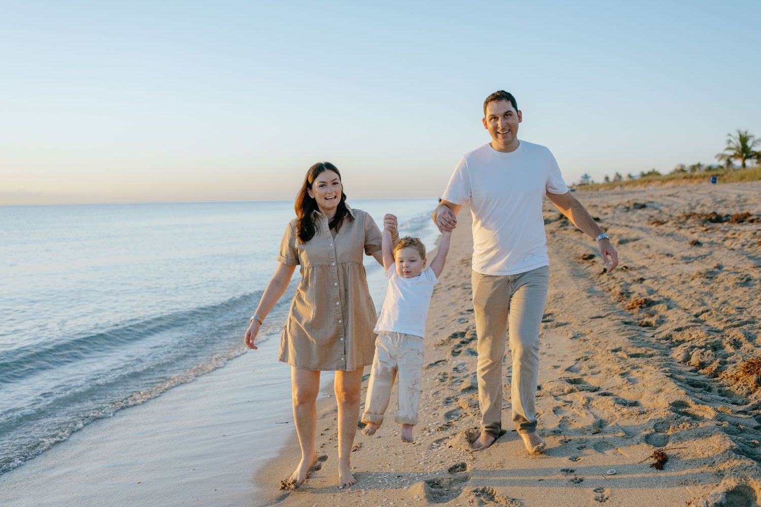 Happy family of three swings their child between them while walking on a beach.