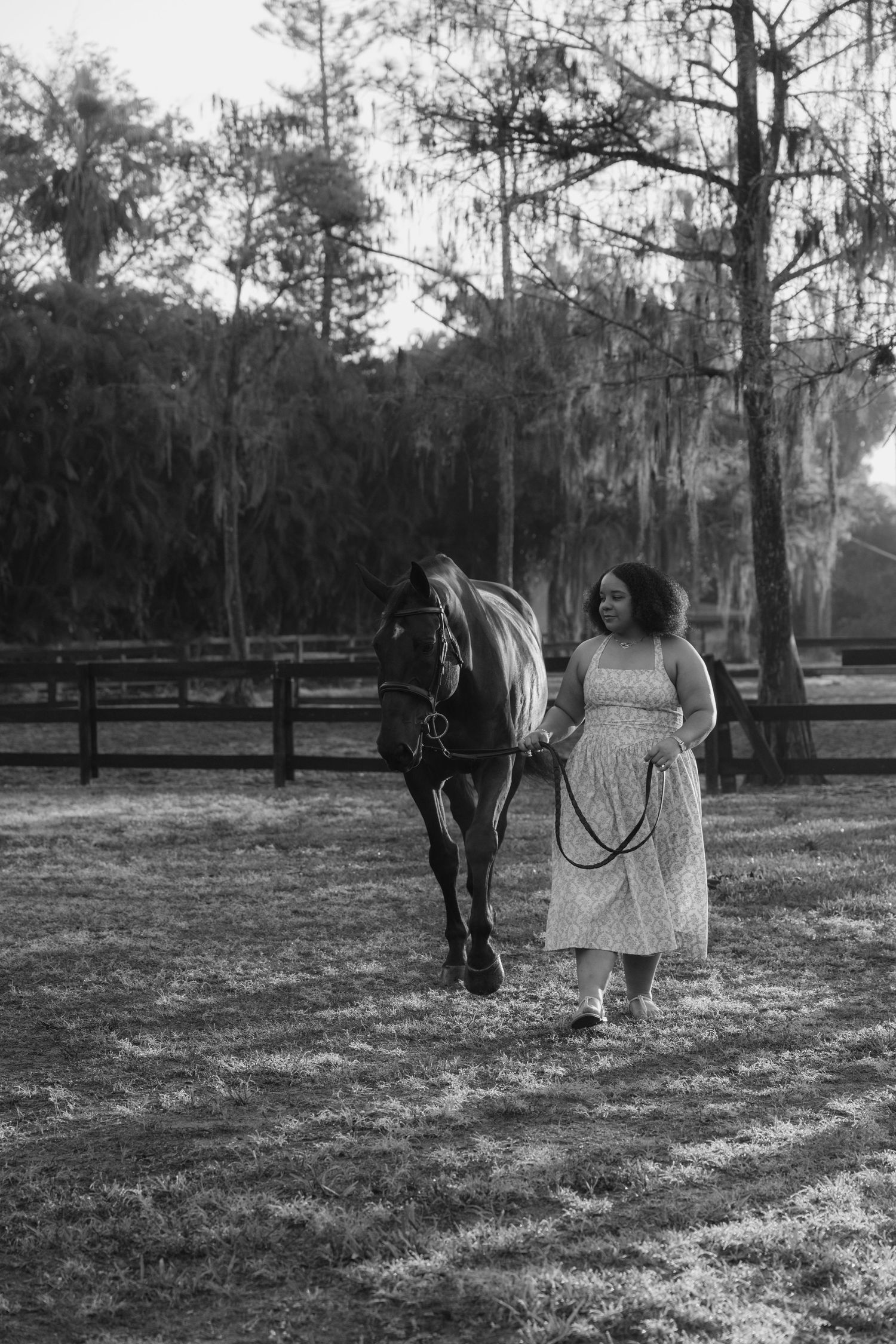 Black and white photo of a woman in a floral dress walking a dark horse in a sunny paddock.