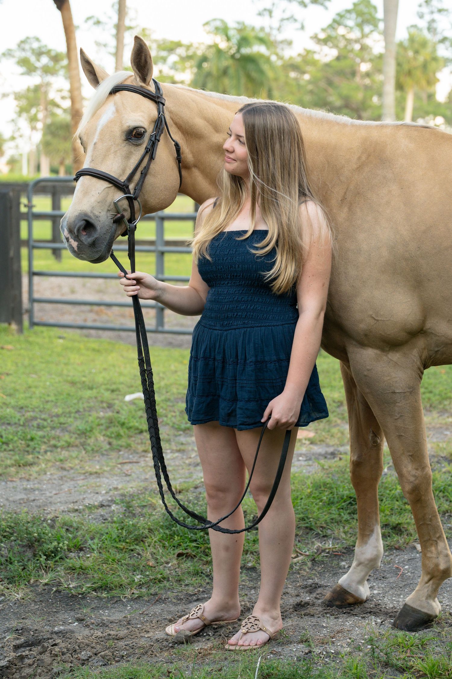 Smiling young woman in navy smocked dress holds lead rope of palomino horse in grassy paddock area.