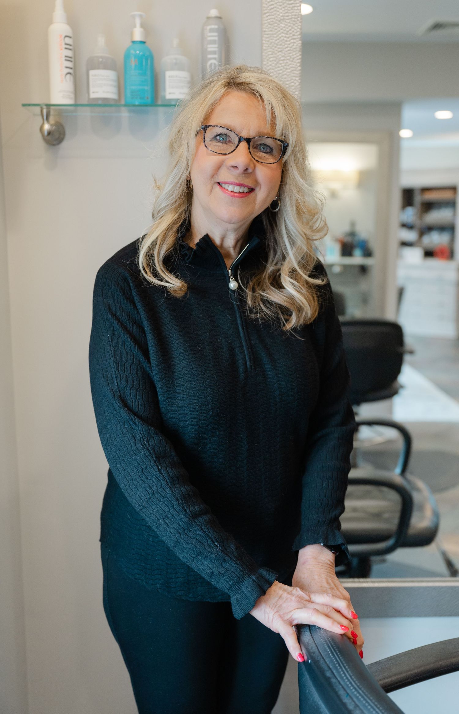 Smiling blonde woman in black outfit posing in modern hair salon with Moroccanoil products on shelf.