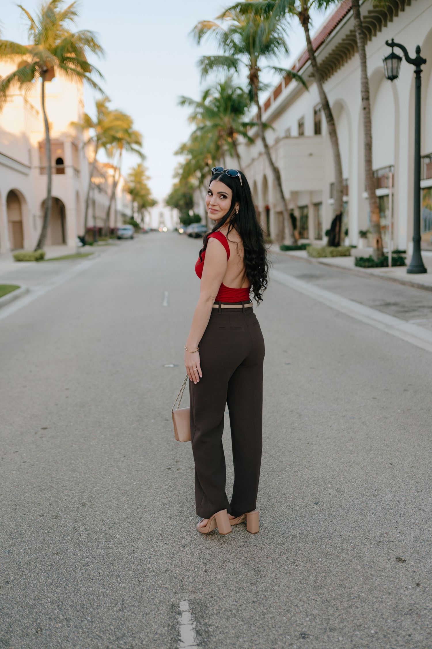 Woman in red top and dark trousers posing on palm-lined street holding beige bag.