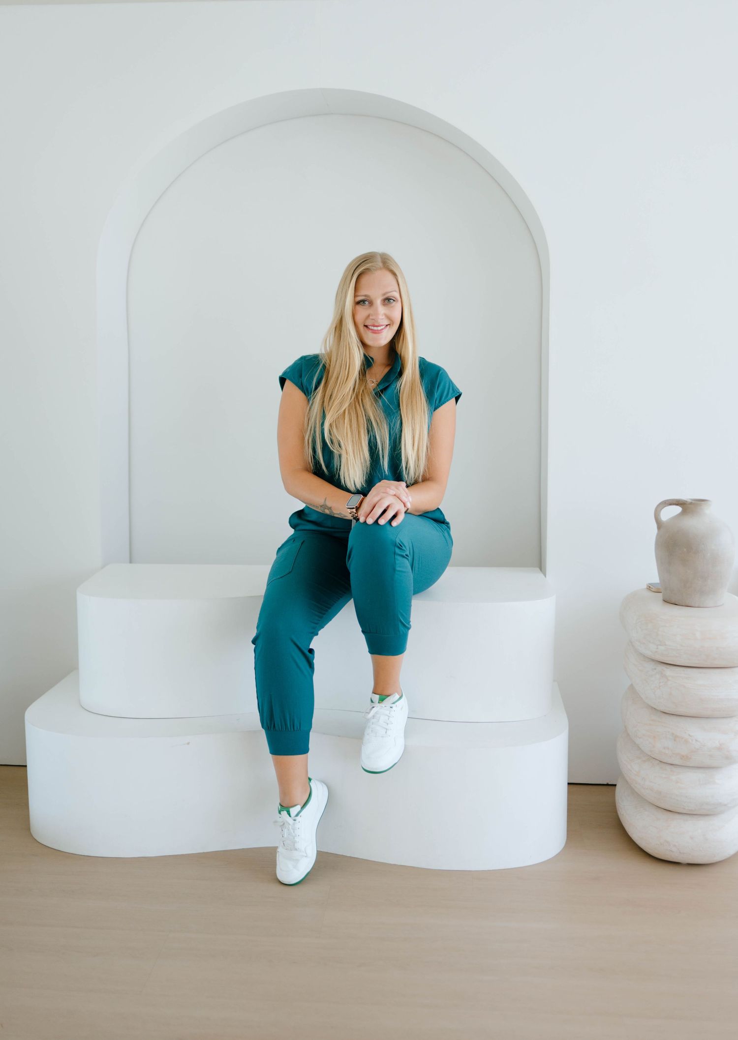 Confident blonde woman in teal scrubs seated on a white tiered platform inside an arched alcove with decor.