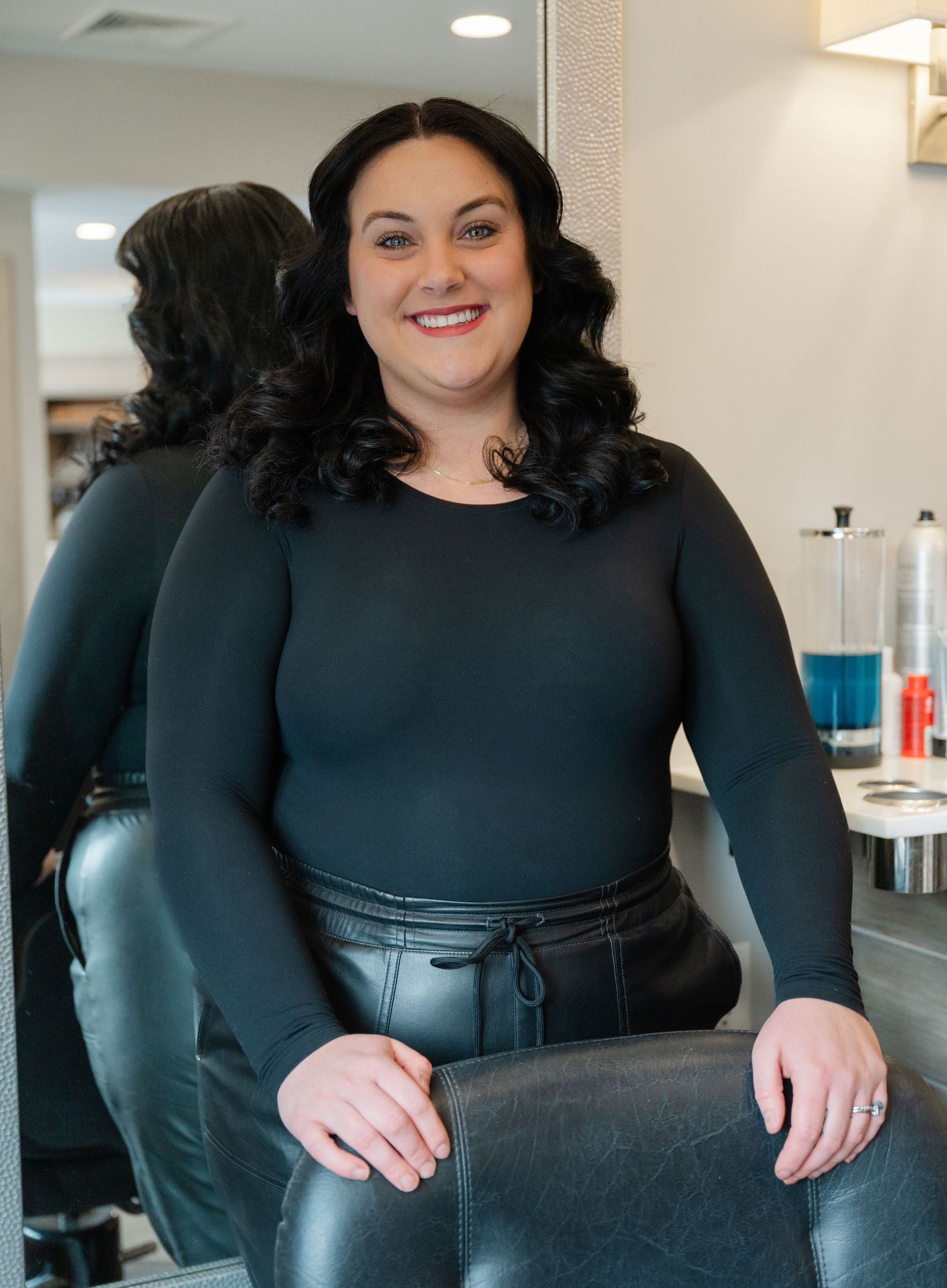 Smiling brunette woman in black bodysuit seated in a hair salon styling chair.