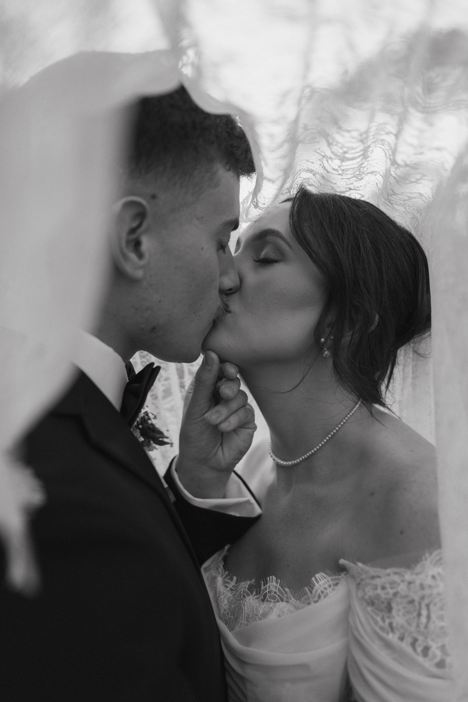 Black and white photo of bride and groom sharing a romantic kiss under a flowing wedding veil.