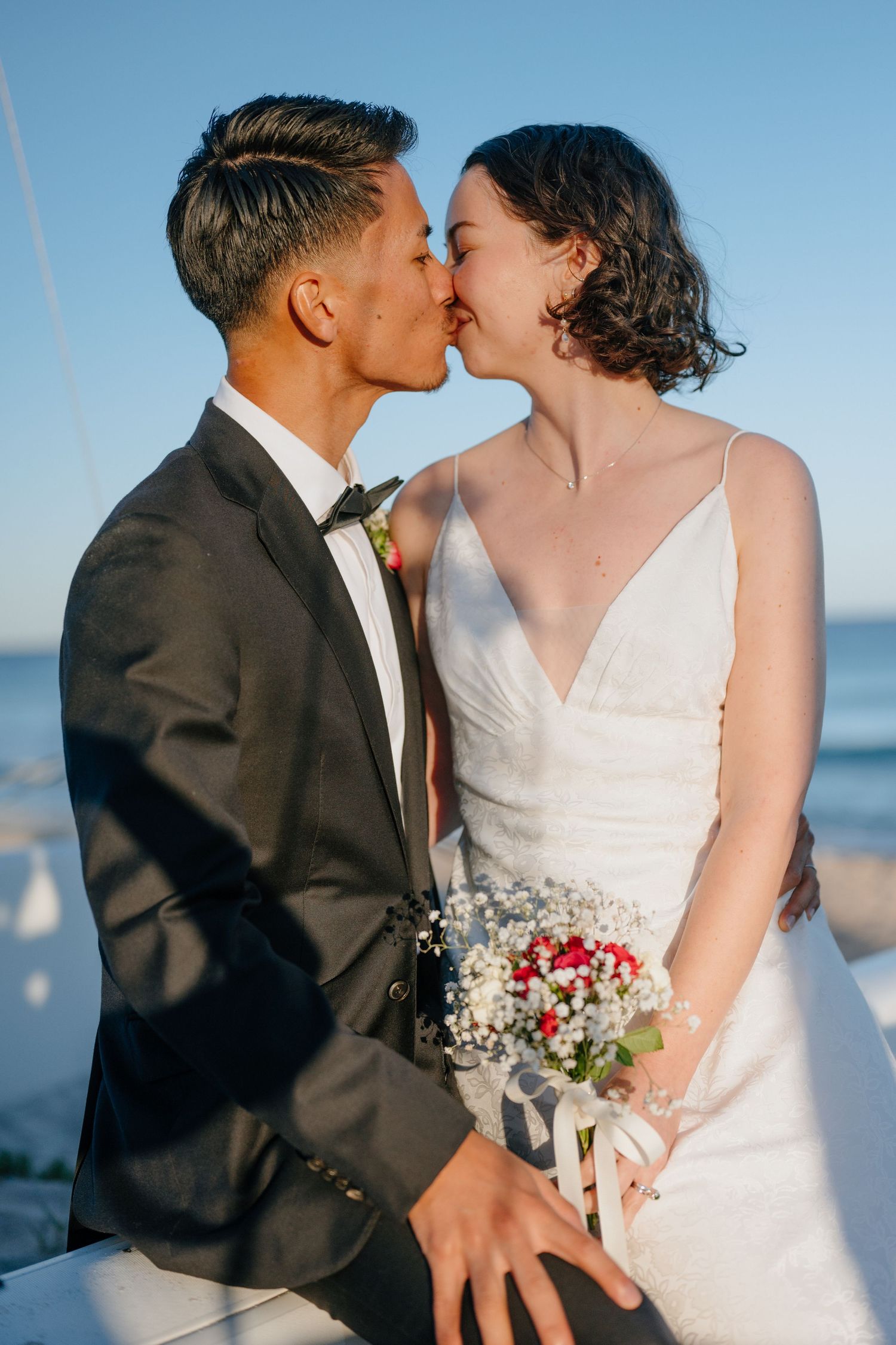 Interracial couple sharing a kiss in wedding attire on a sunny beach, bride holding red and white bouquet.