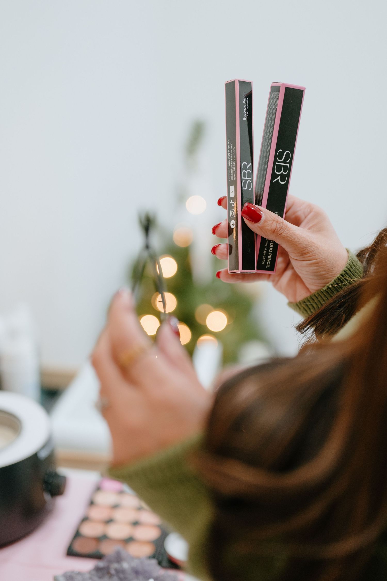 Hands holding two SBR branded pink cosmetic product boxes near a Christmas tree and makeup palette.
